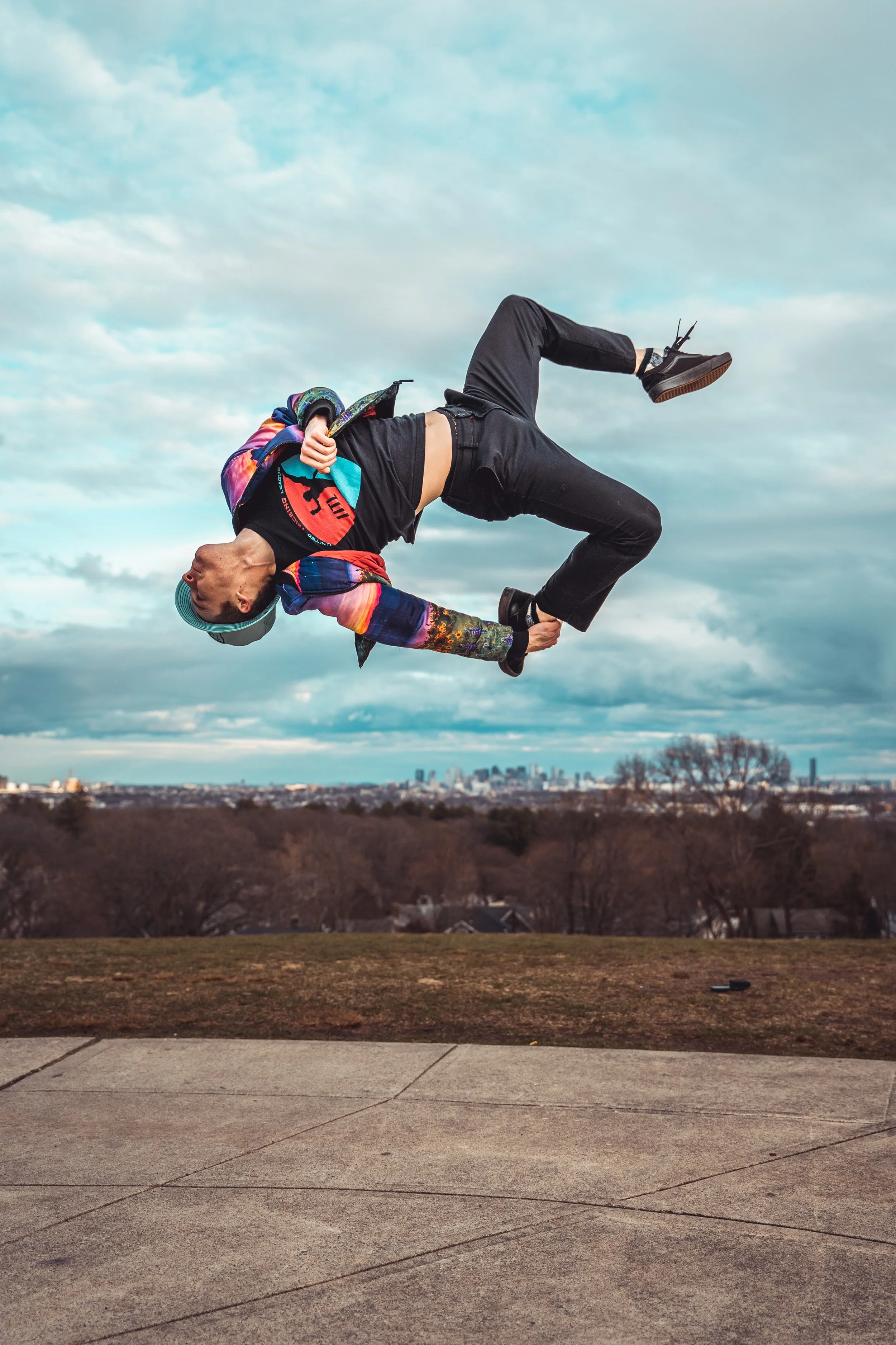 A person performing a backflip outdoors on a cloudy day, with city skyline in the background.