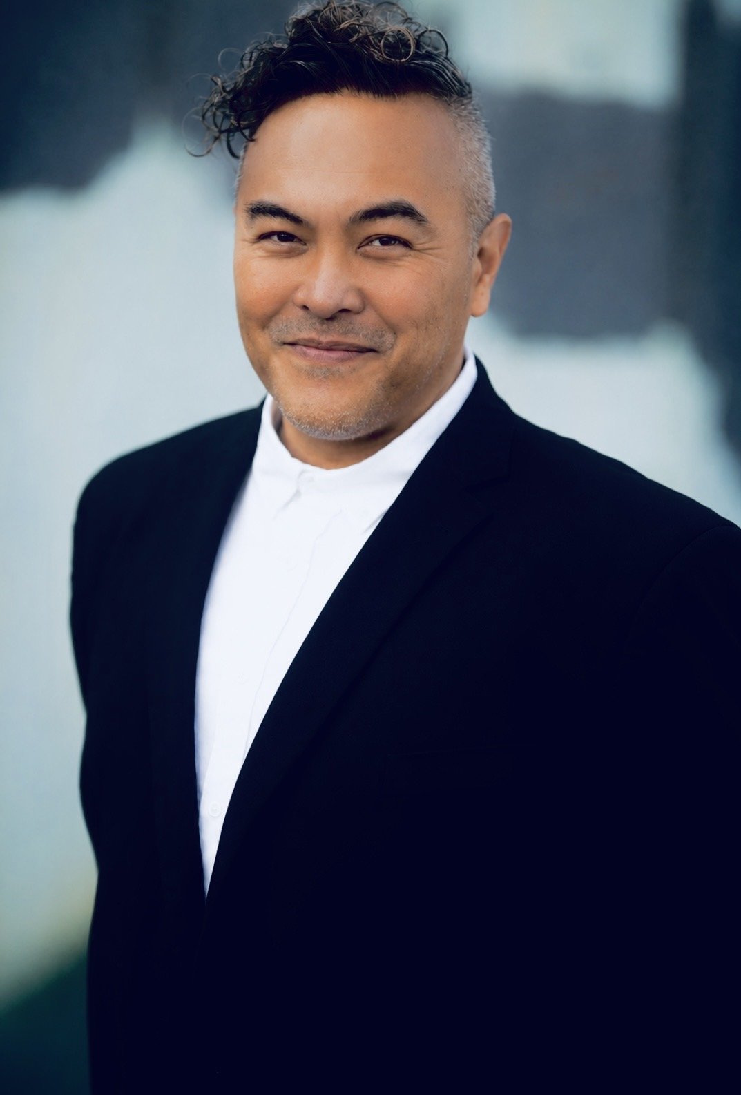 A middle-aged man with short, curly gray hair, wearing a black suit jacket over a white dress shirt, smiling in a professional headshot. The background is blurred with faint colors of green and gray.
