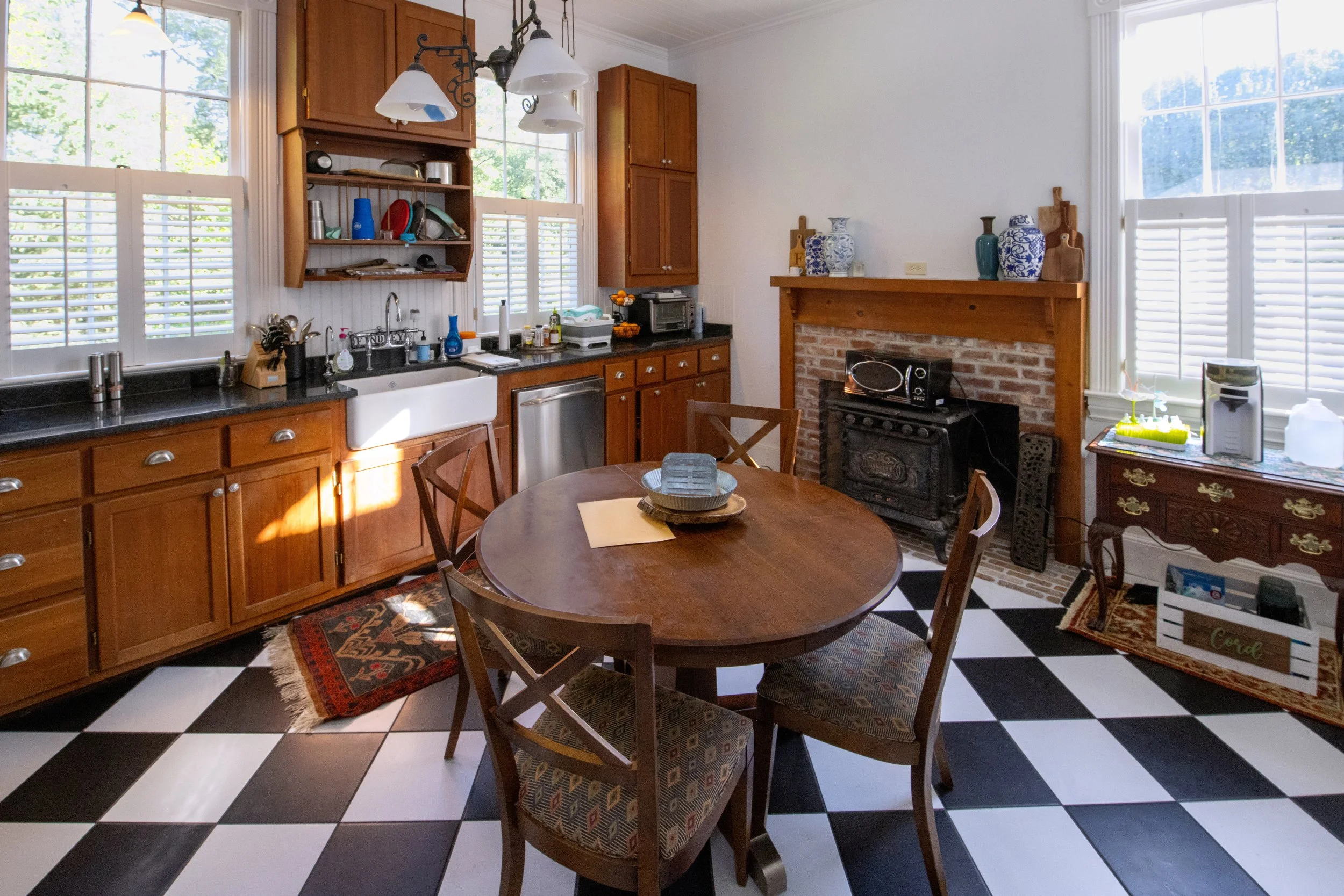 A cozy kitchen with wooden cabinets, checkered black and white floor, round wooden dining table with four chairs, and a brick fireplace mantel decorated with blue and white vases.