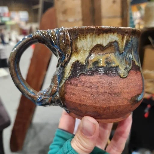 A handmade ceramic mug with a tan, brown, and beige glaze, featuring a leaf design on the side.