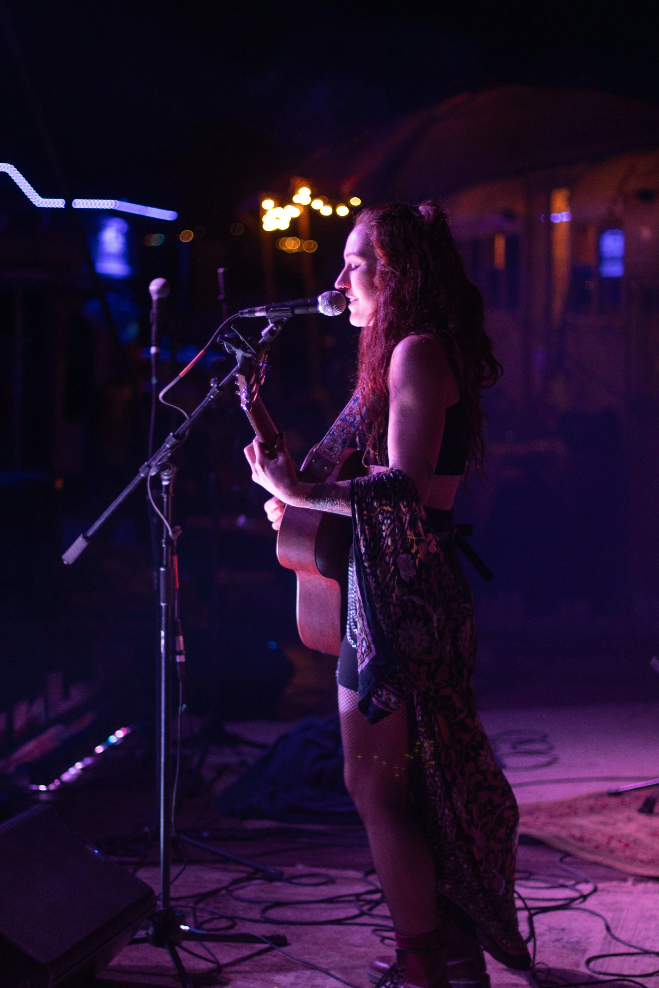 Female musician performing on stage at night, playing guitar and singing into microphone with colorful stage lighting.