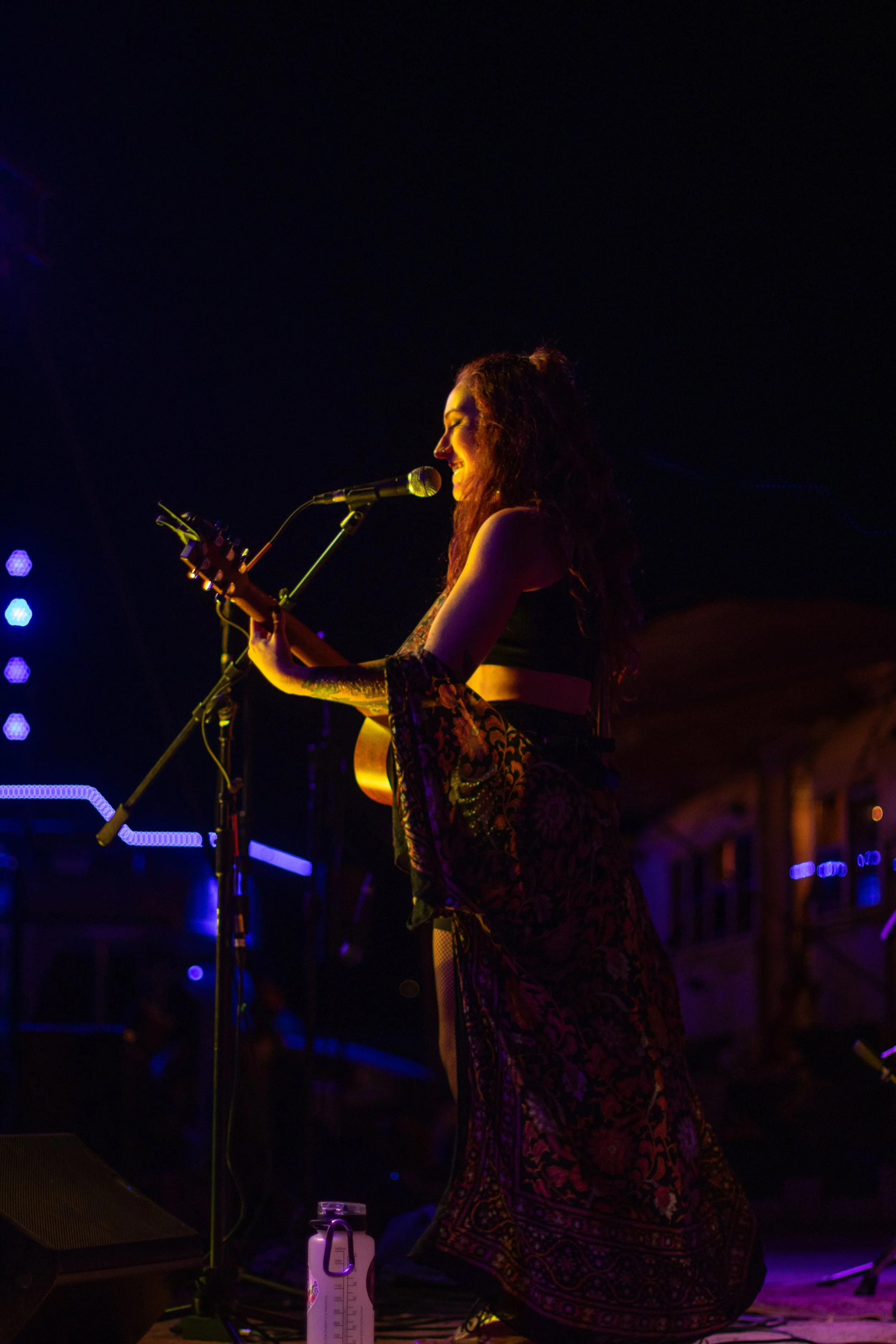 A female musician singing and playing guitar on stage under stage lights, with a dark background.