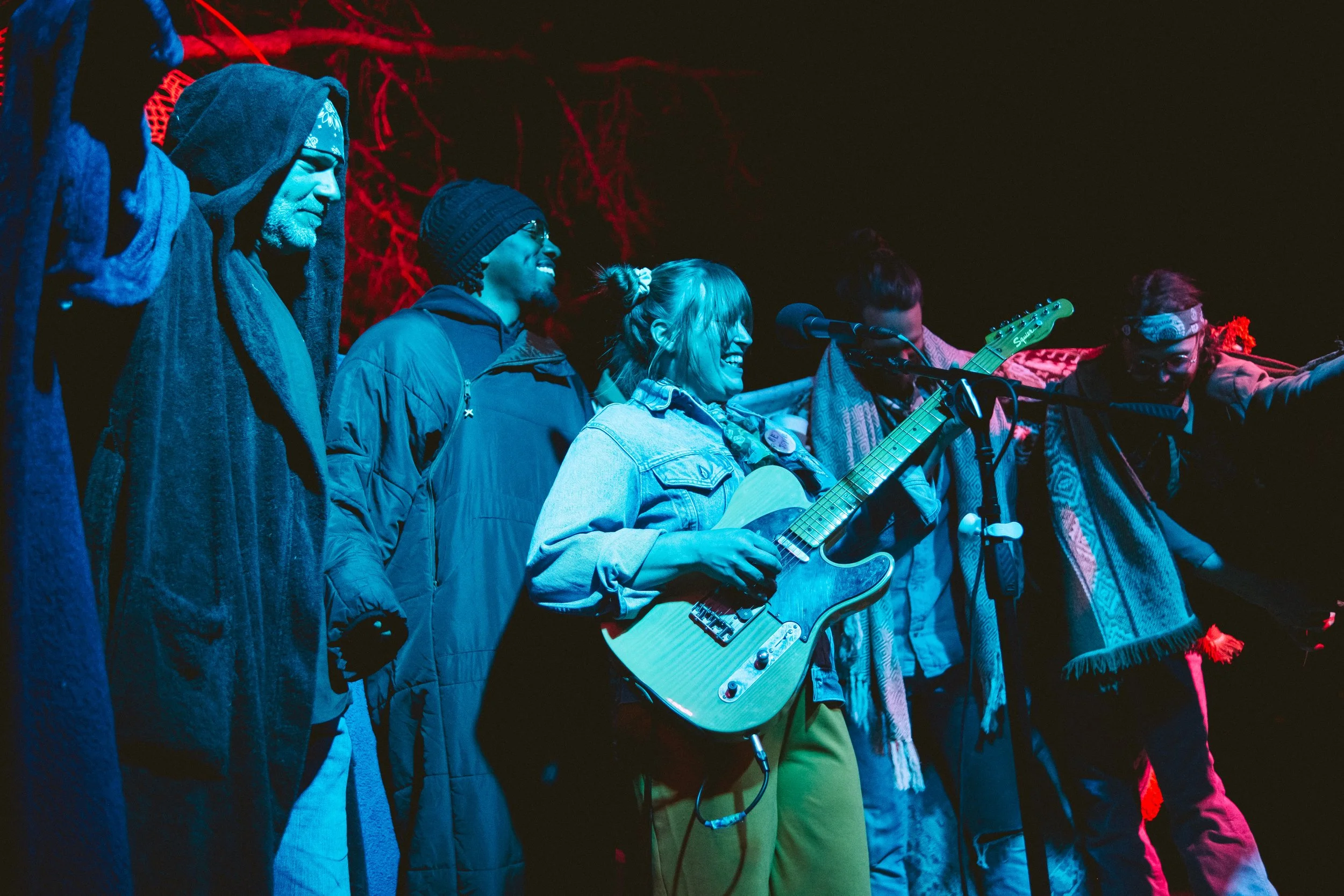 Group of people on stage at night, with one woman playing guitar and singing into a microphone, others cheering and clapping, illuminated by colorful stage lights