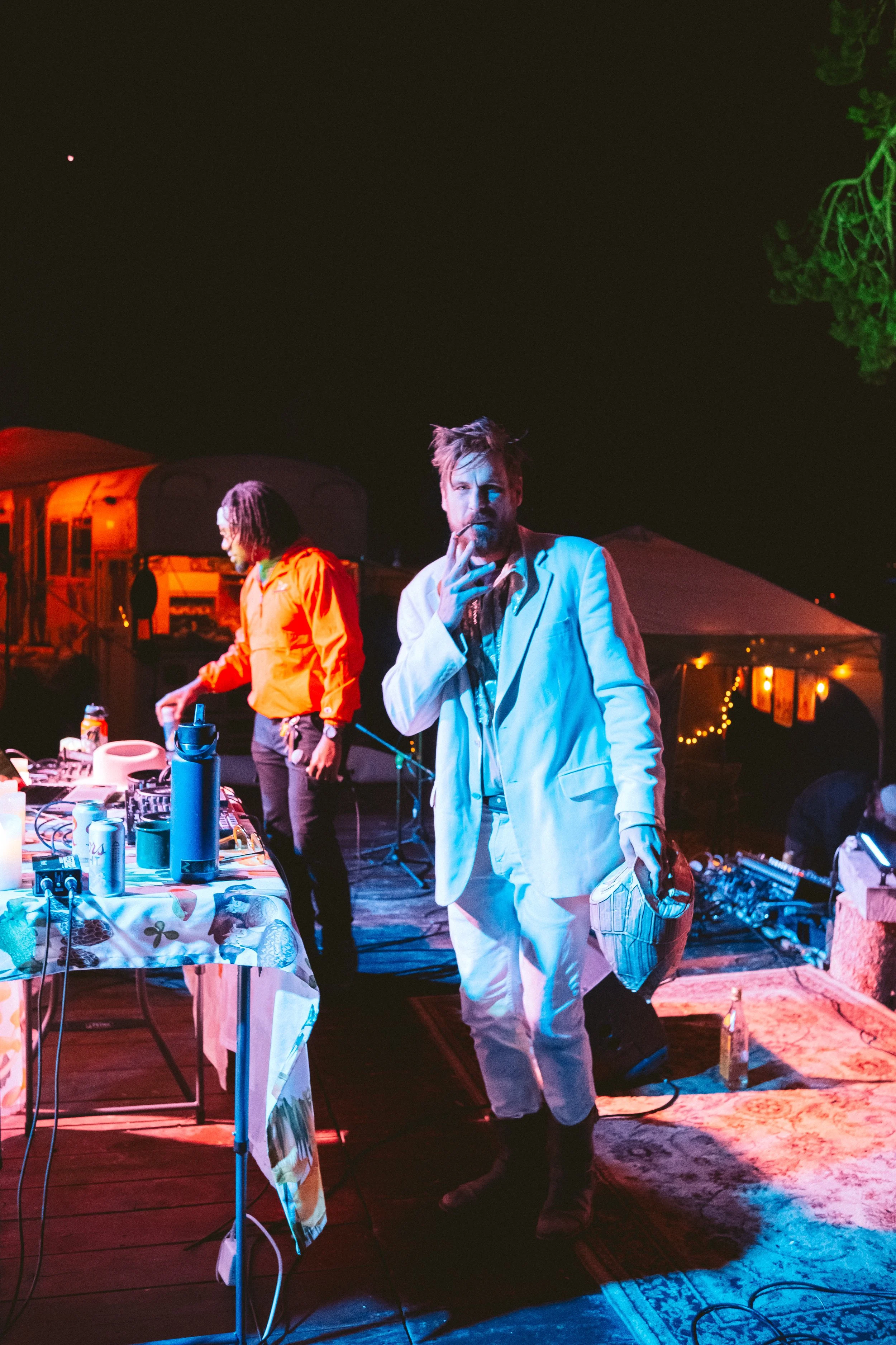 Man in a white suit with a cigarette in his hand, standing near a table with DJ equipment and drinks at an outdoor nighttime event.