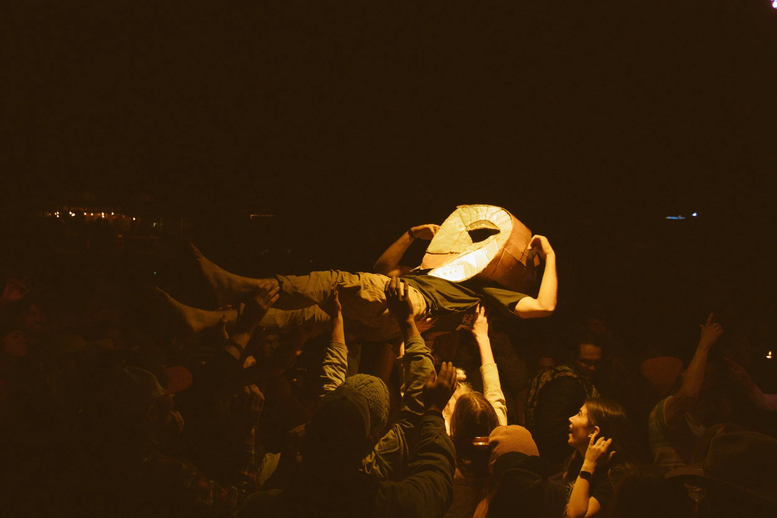 People gathered around a bonfire at night, holding a person up wearing a large paper lantern mask in the shape of a smiling face.