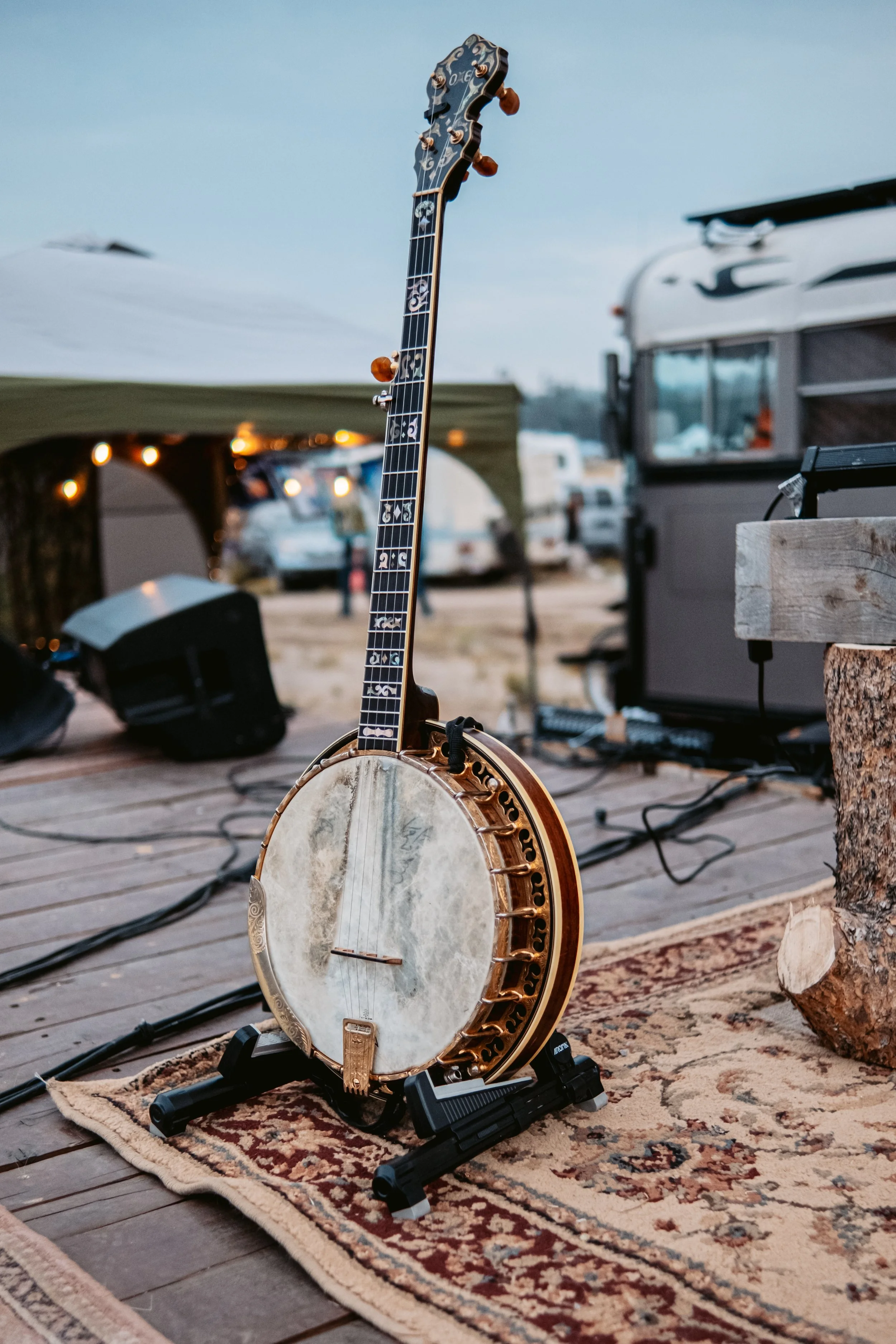 A banjo with a guitar neck, placed on a stand on a rug on a stage outdoors, with a backdrop of tents, RVs, and light strings at dusk.