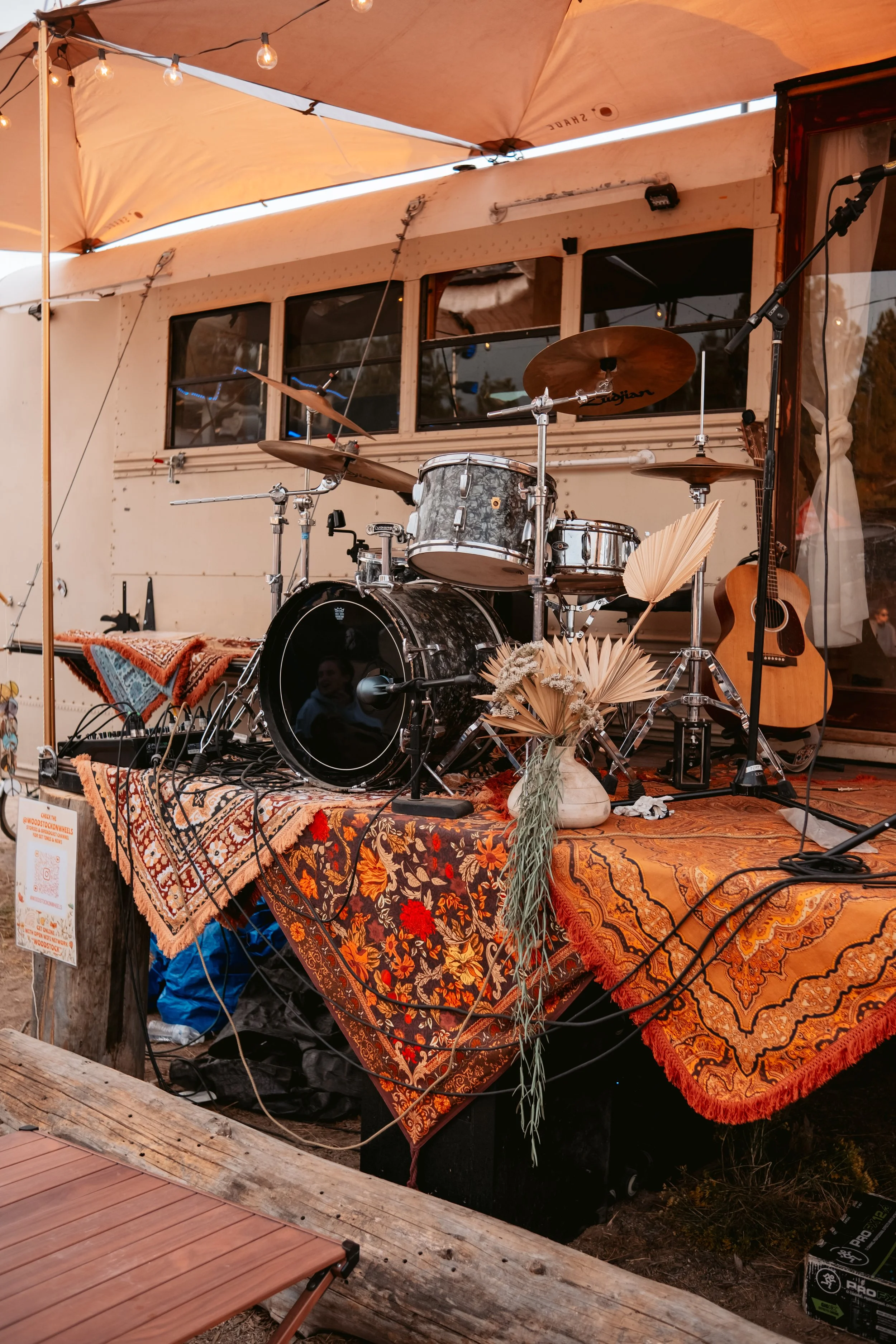 Stage with drums, guitar, and decorated table with dried flowers under a tent at an outdoor event.