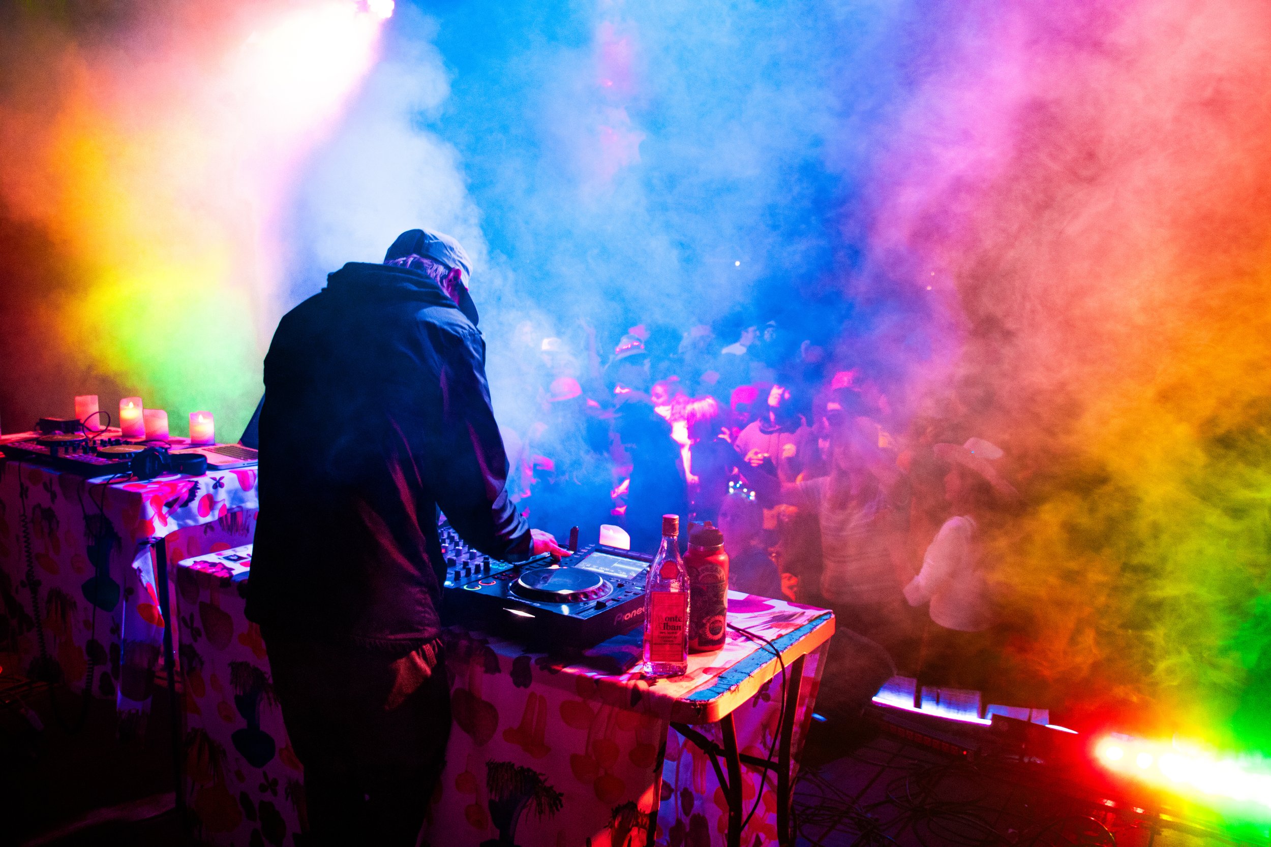 DJ performing at a colorful nightclub with a crowd dancing in the background, smoke effects, and vibrant lighting.
