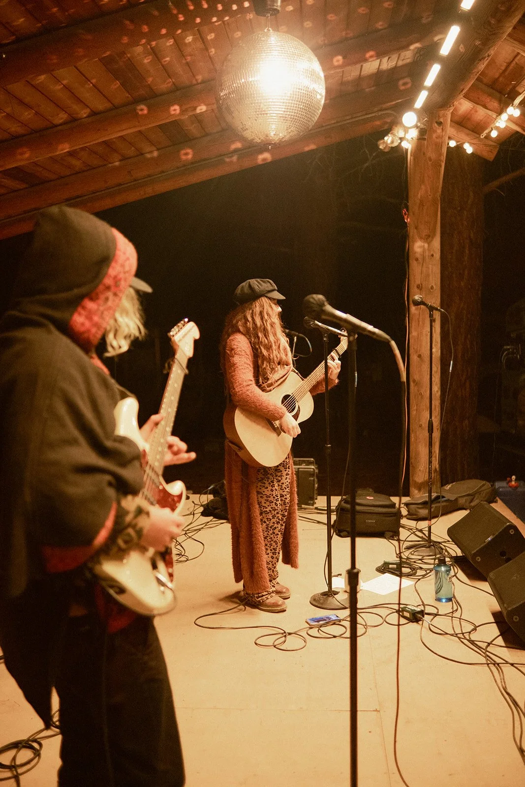 Three musicians performing on stage with guitars, microphones, and equipment at a rustic venue with wooden beams and a disco ball.
