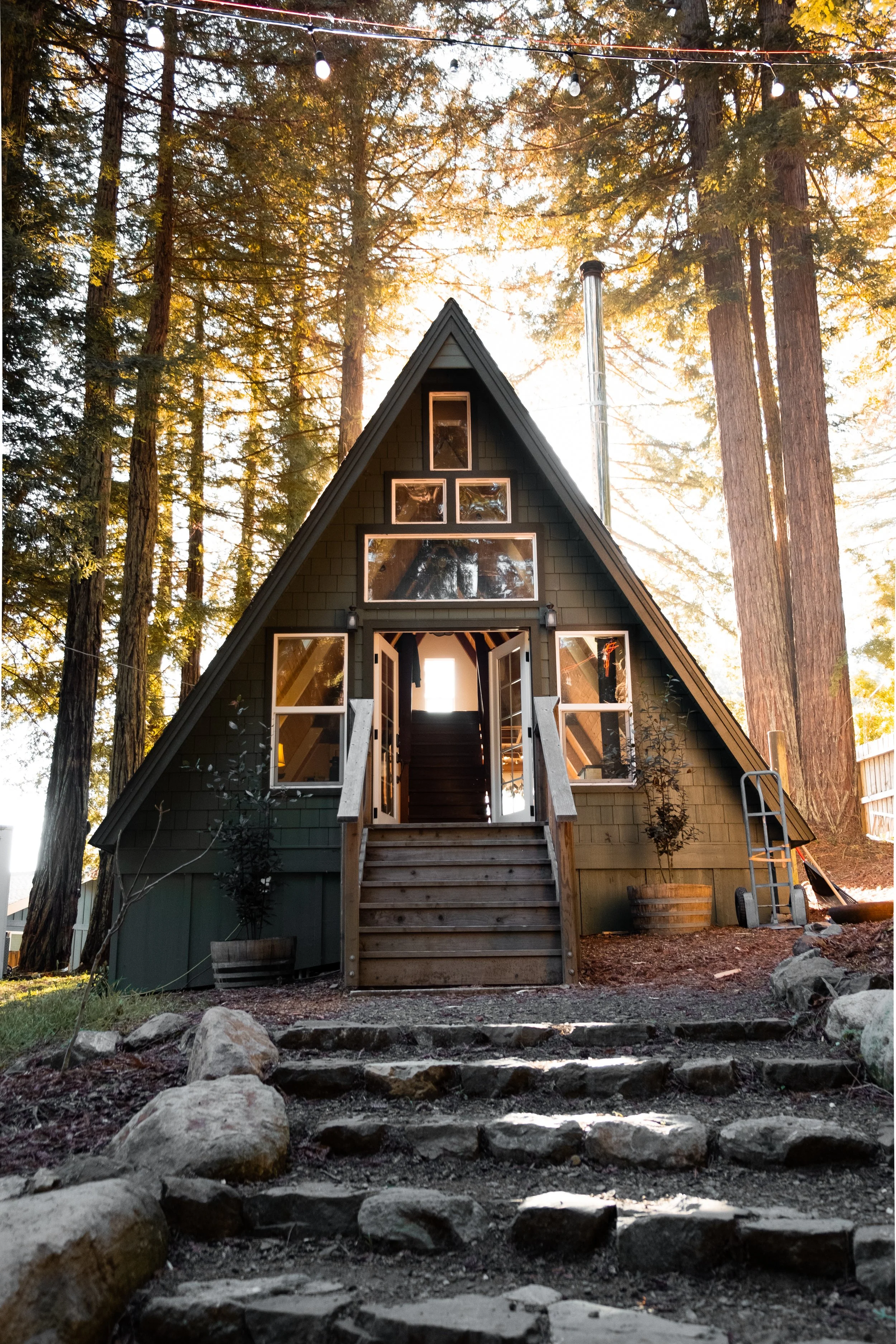 A small black A-frame house with open front doors, surrounded by tall trees, with sunlight shining through the branches.