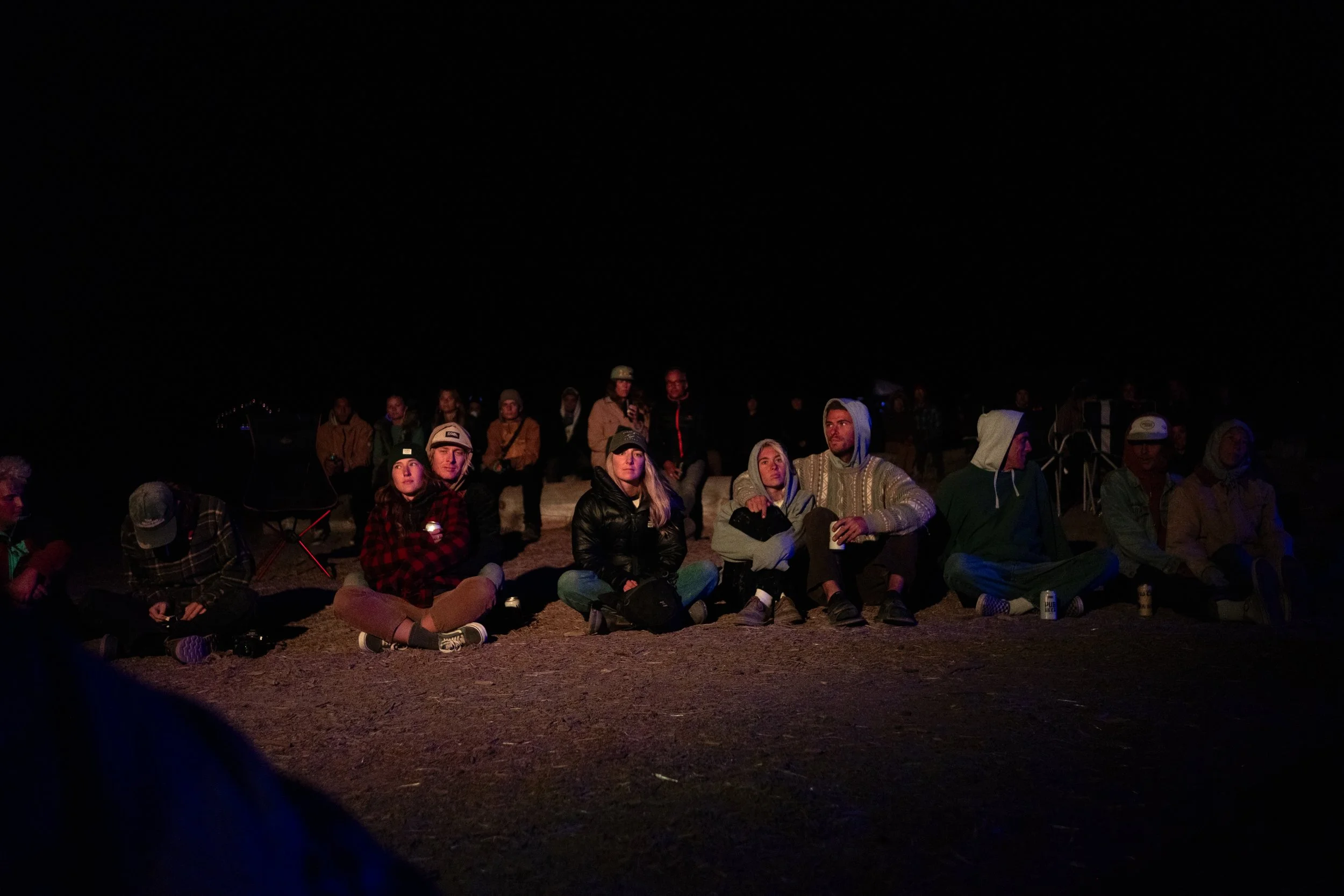 A group of people sitting and watching at night, some holding drinks, with a dark sky above.