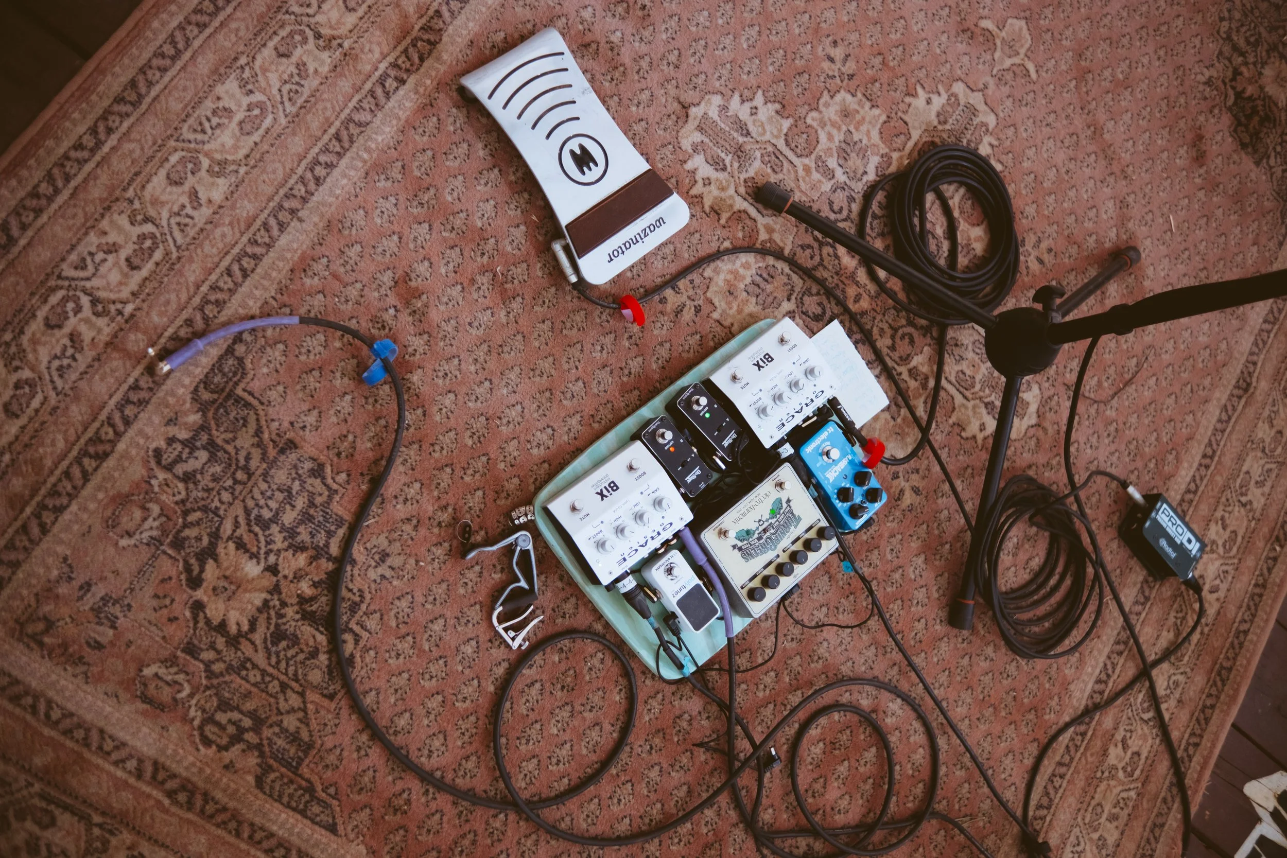 Audio recording equipment including multiple guitar effect pedals, cables, a microphone stand, and a wireless transmitter on a patterned carpet.
