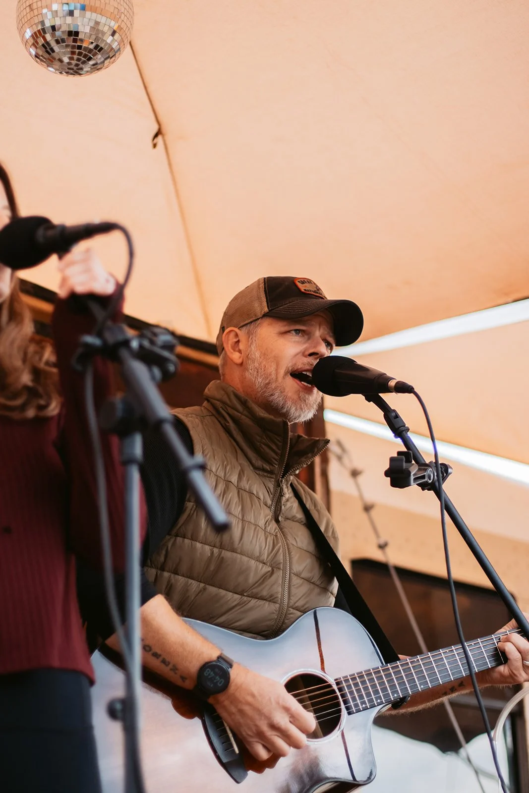 A man singing and playing an acoustic guitar on stage, with a microphone in front of him, under a beige canopy, with part of a disco ball visible in the corner.