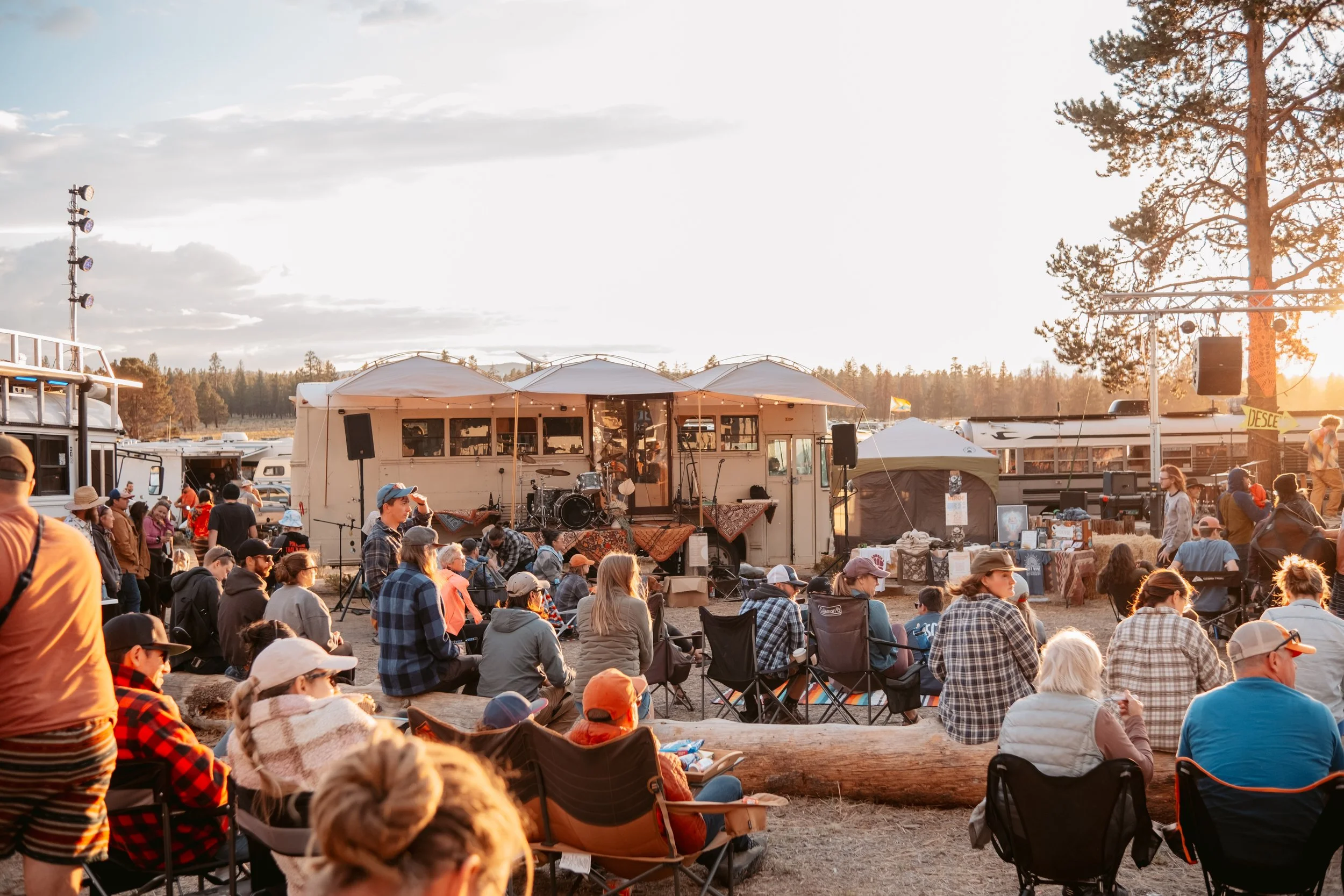 Outdoor concert scene with people sitting on chairs and logs watching a band perform on stage at sunset, with boats and trees in the background.
