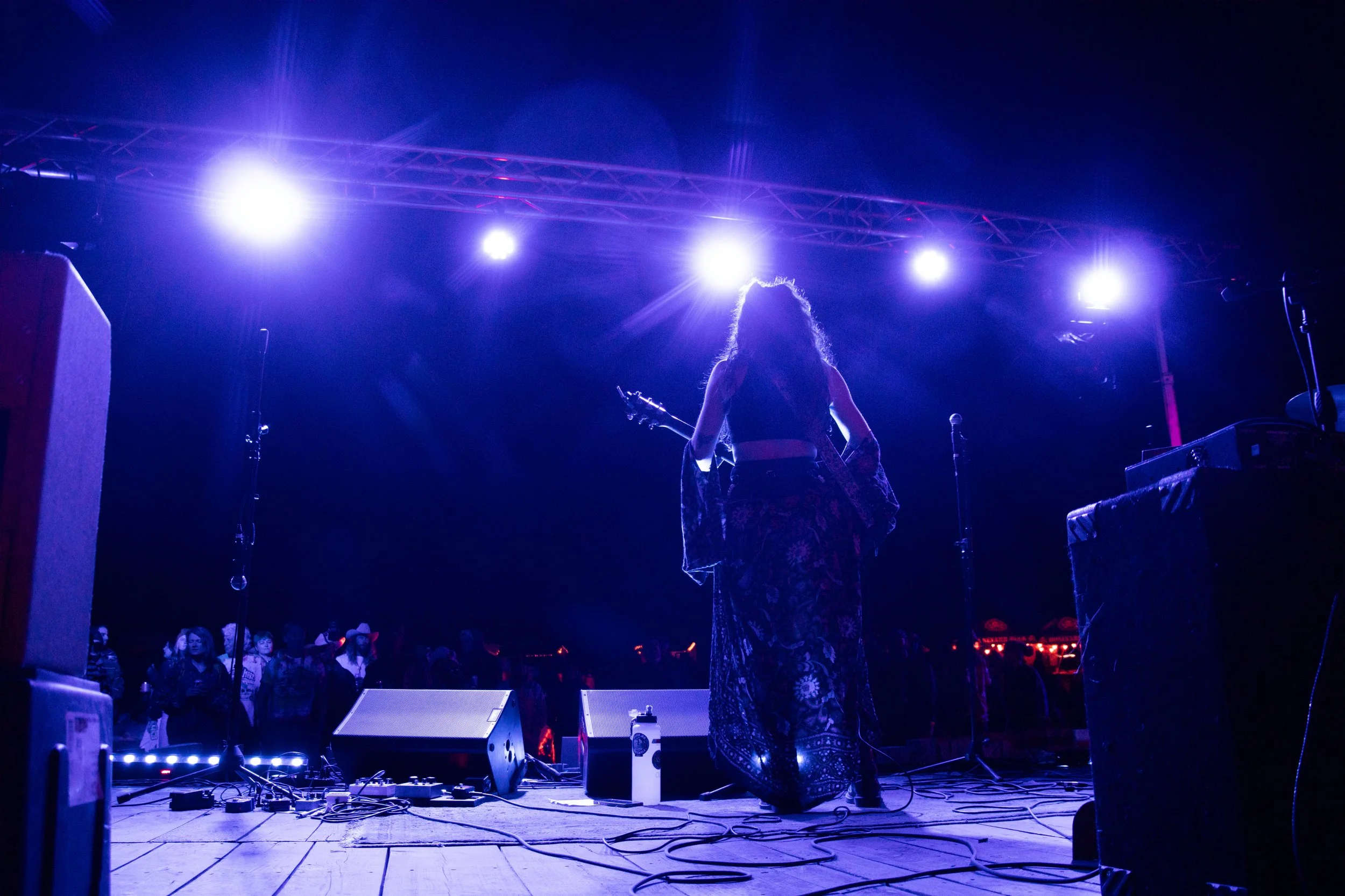 A female musician on stage playing guitar at night, backlit by bright purple and white stage lights, with an audience in the background.