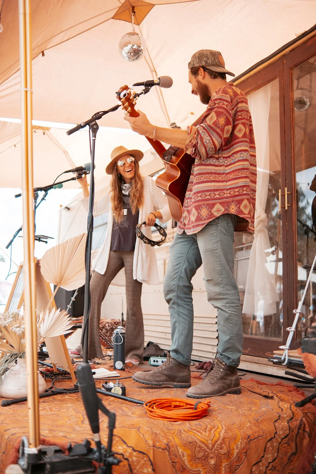 A man playing guitar and singing into a microphone, smiling woman with sunglasses and a hat holding a tambourine, outside under a large beige umbrella with a disco ball hanging above, on a decorated patio with orange carpet and plants.