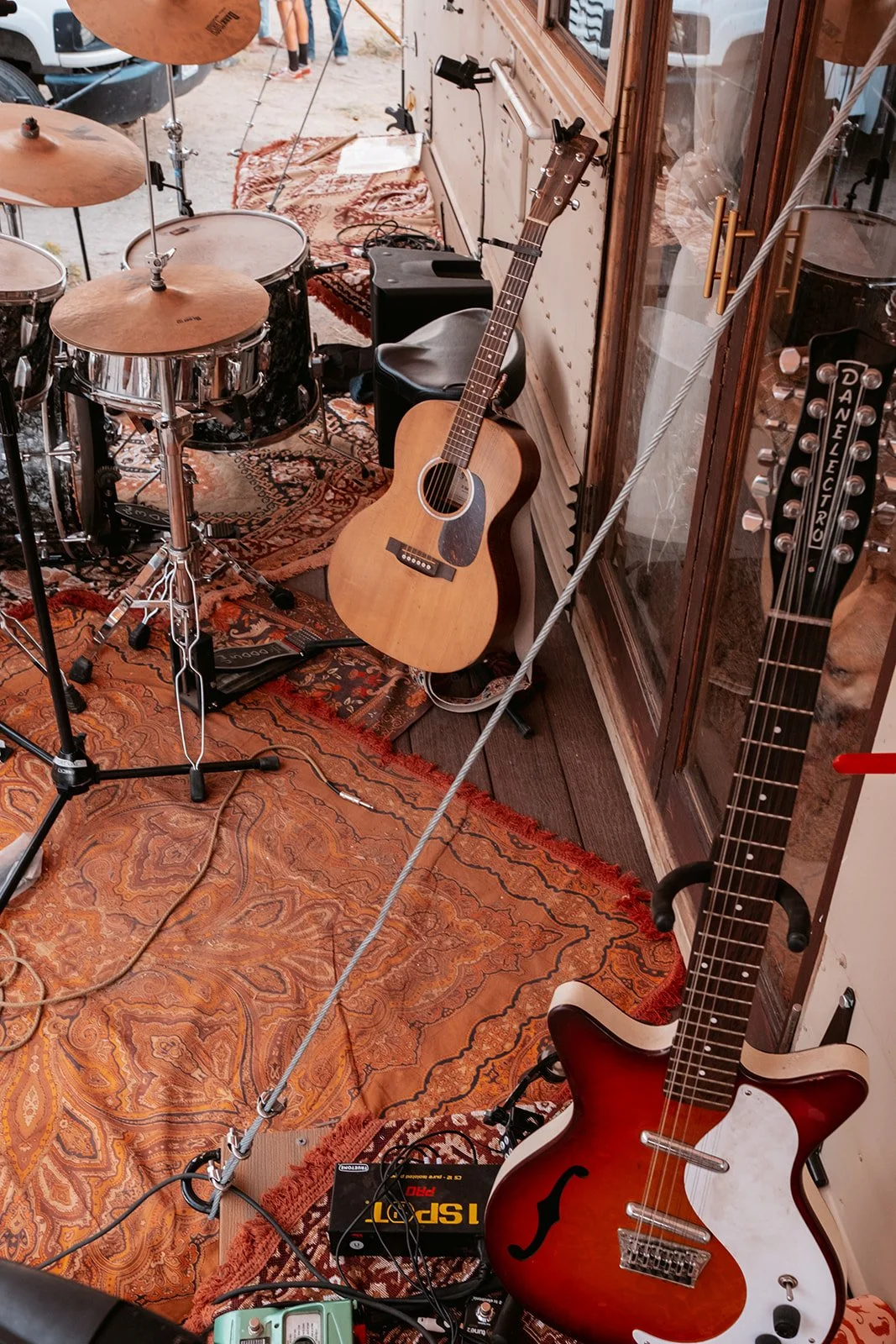 Musical instruments including a drum set, an acoustic guitar, and an electric guitar are set up on a patterned rug in a room or studio.