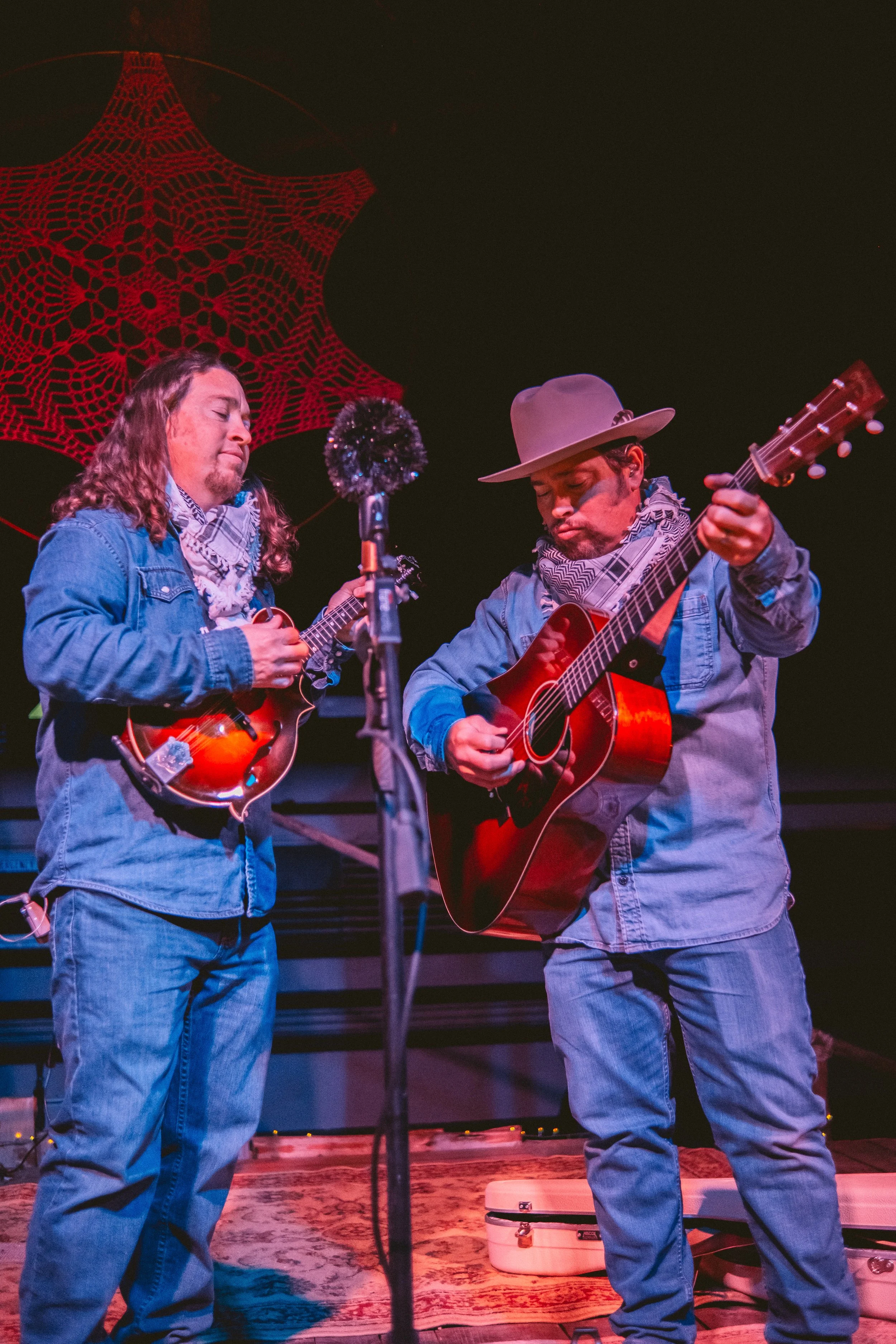 Two men wearing denim shirts and scarves playing acoustic guitars on stage, with a red web-like decoration in the background.