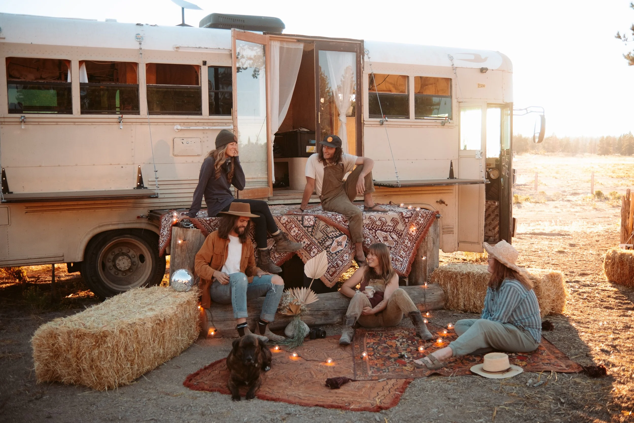 Group of people relaxing outdoors near an old camper van at sunset, sitting on hay bales and rugs with string lights, some wearing hats and casual clothes, with a small dog in the foreground.