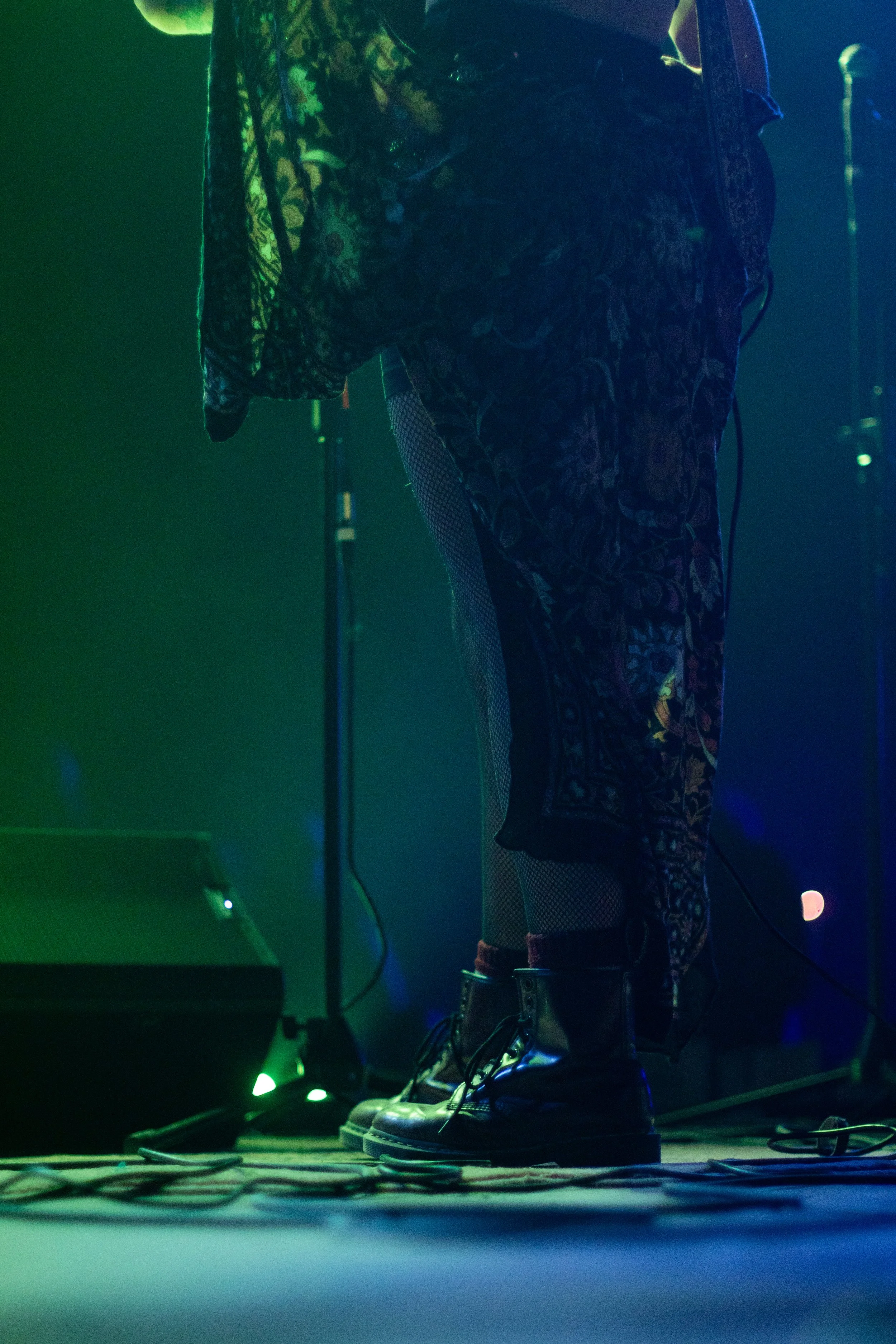 Close-up of a person's lower body on stage, wearing patterned pants, fishnet socks, and black boots under green and blue stage lighting.
