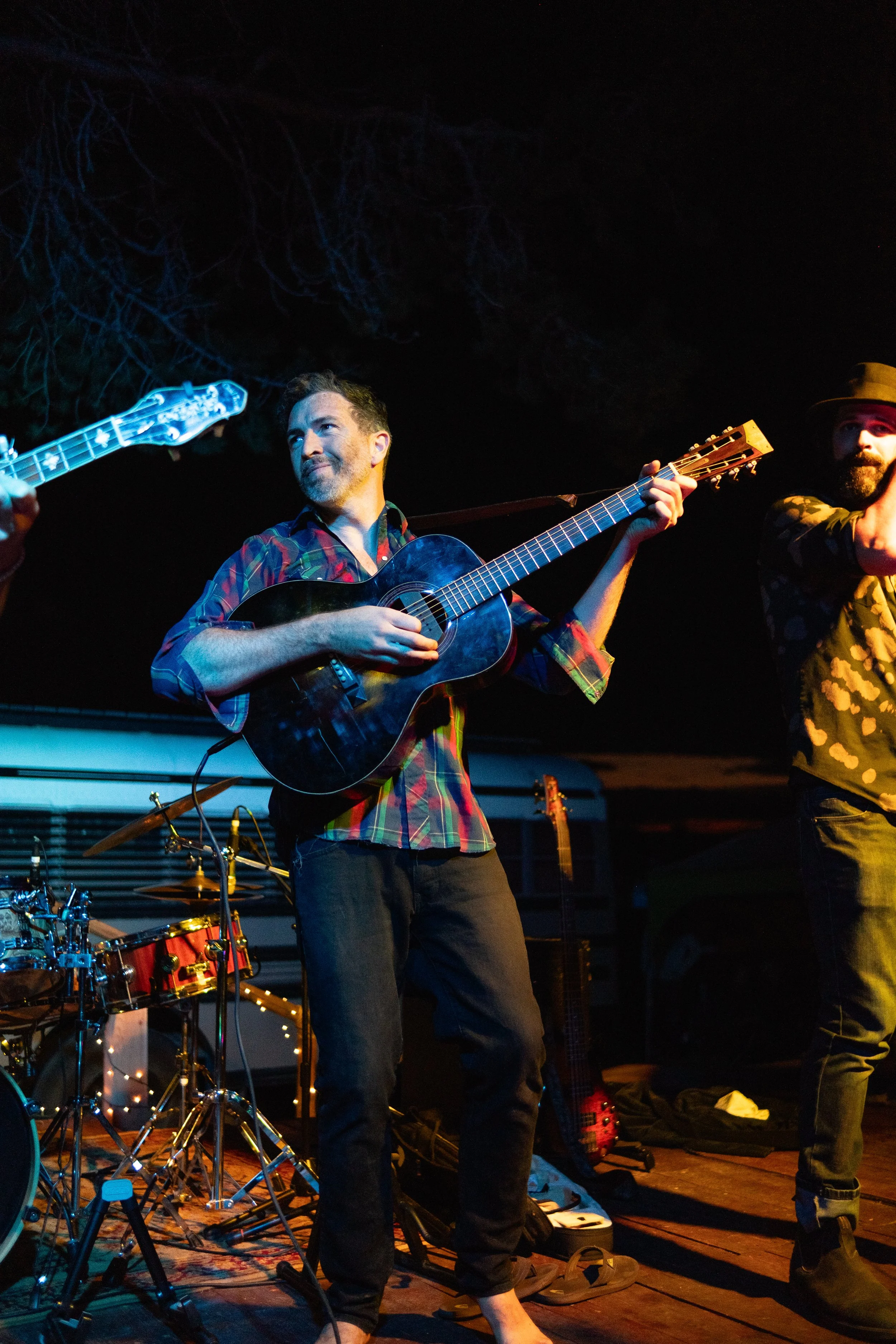 Musicians performing outdoors at night, one playing acoustic guitar, with drums and other instruments in the background.