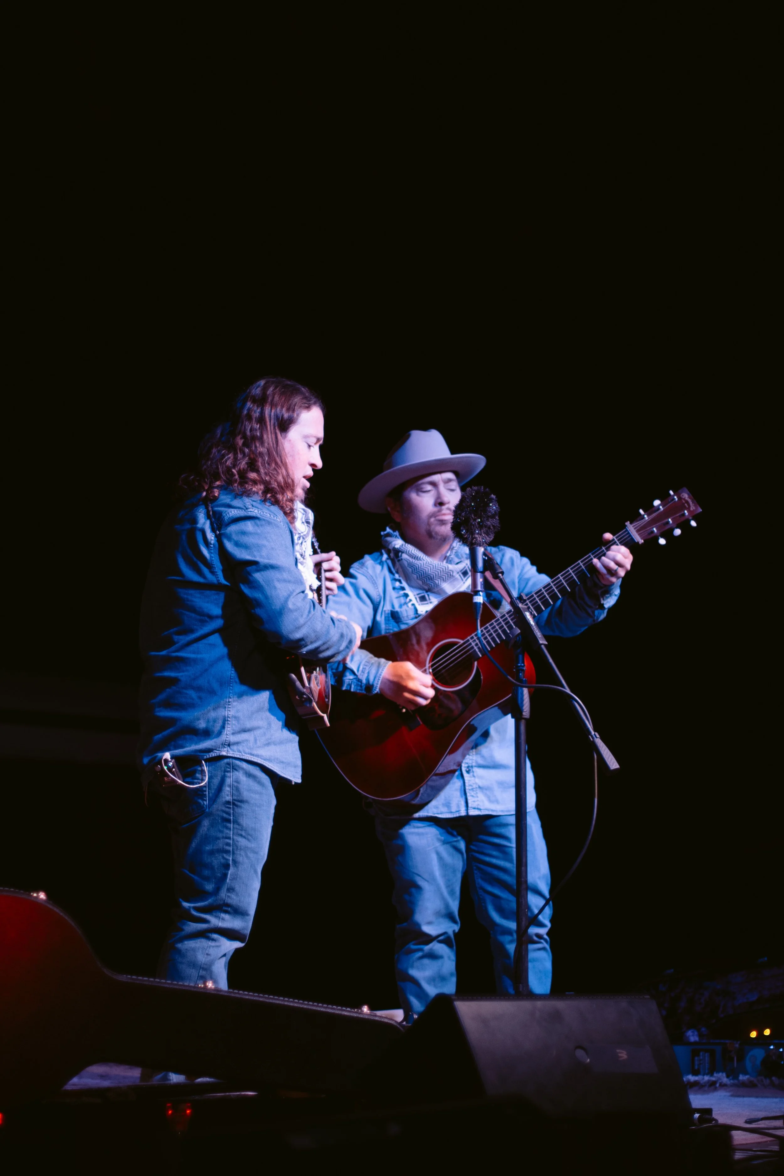 Two musicians perform on stage with guitars, one singing into a microphone, against a dark background.