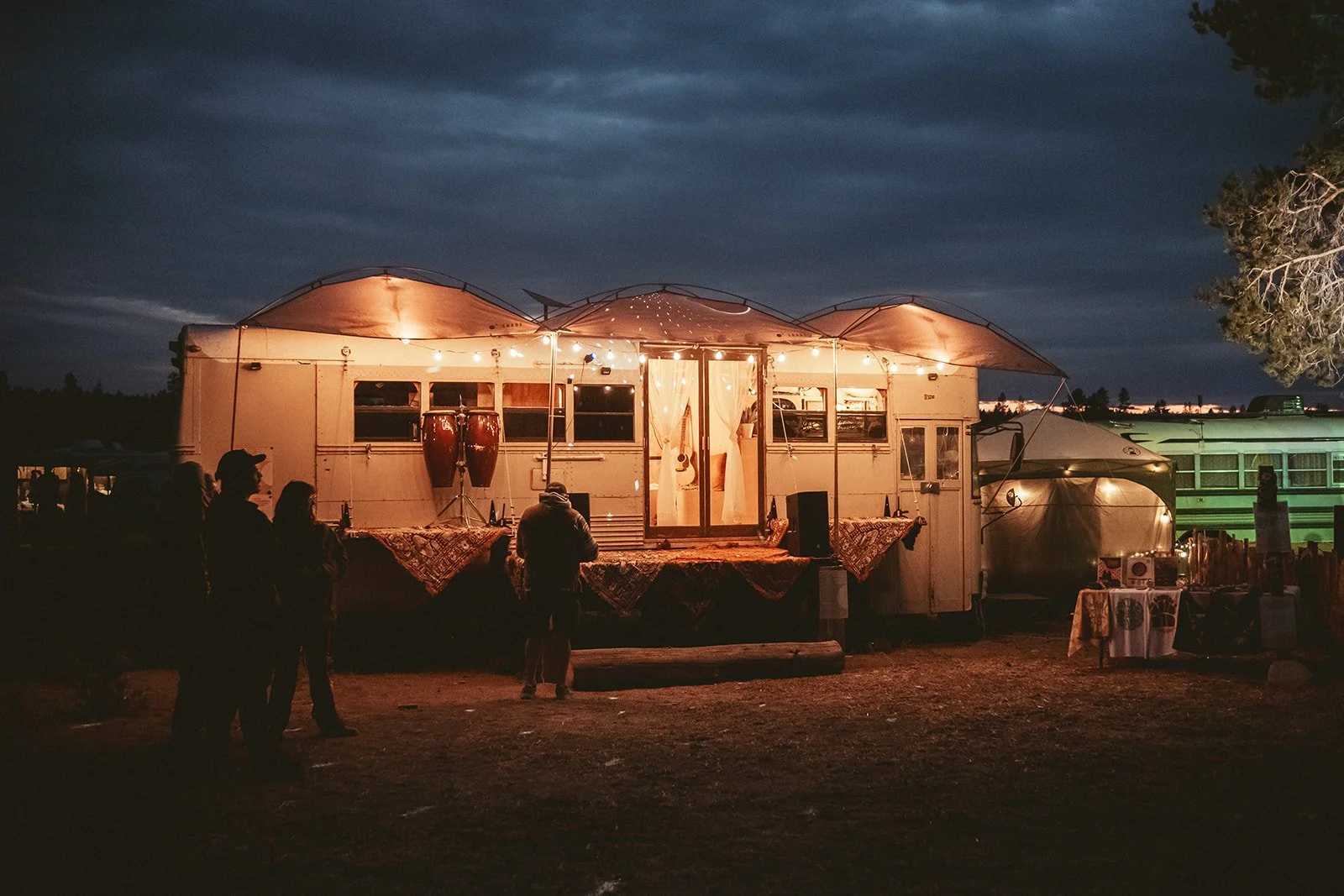 A vintage camper trailer decorated with string lights and hanging drums at night, with people standing nearby.