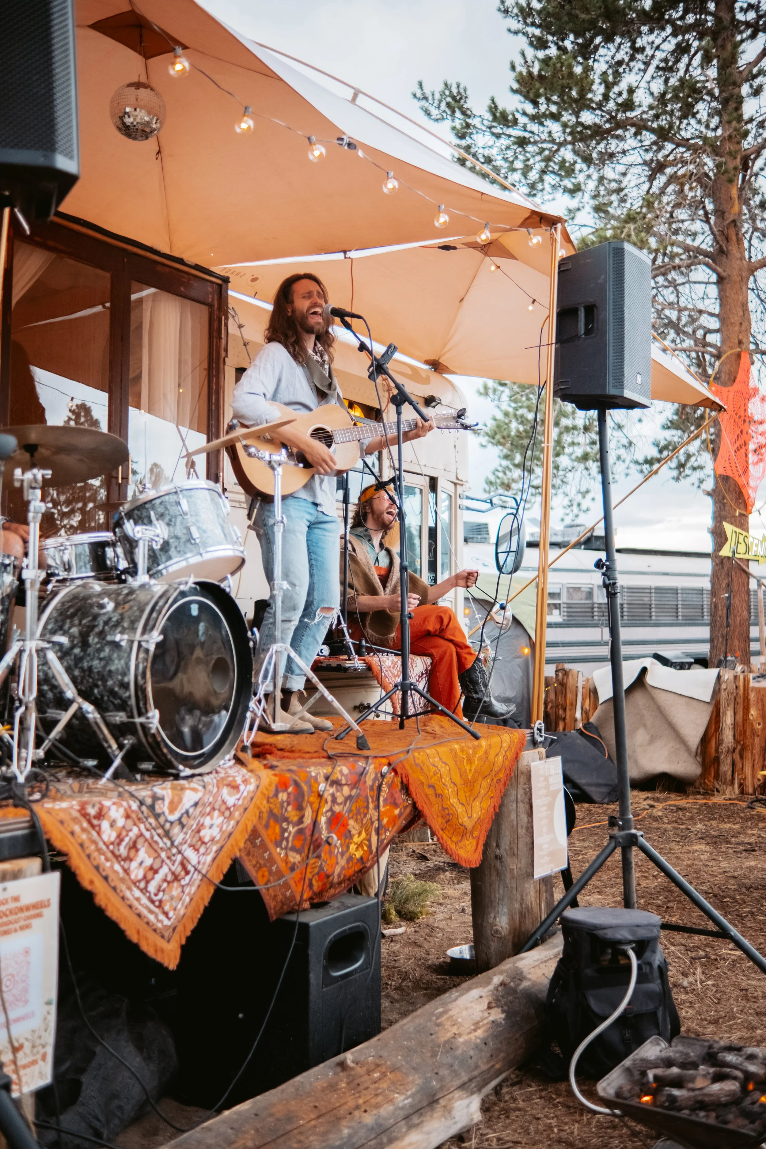 A musician playing an acoustic guitar and singing on an outdoor stage with a band, during sunset with string lights overhead, in a rustic outdoor setting.