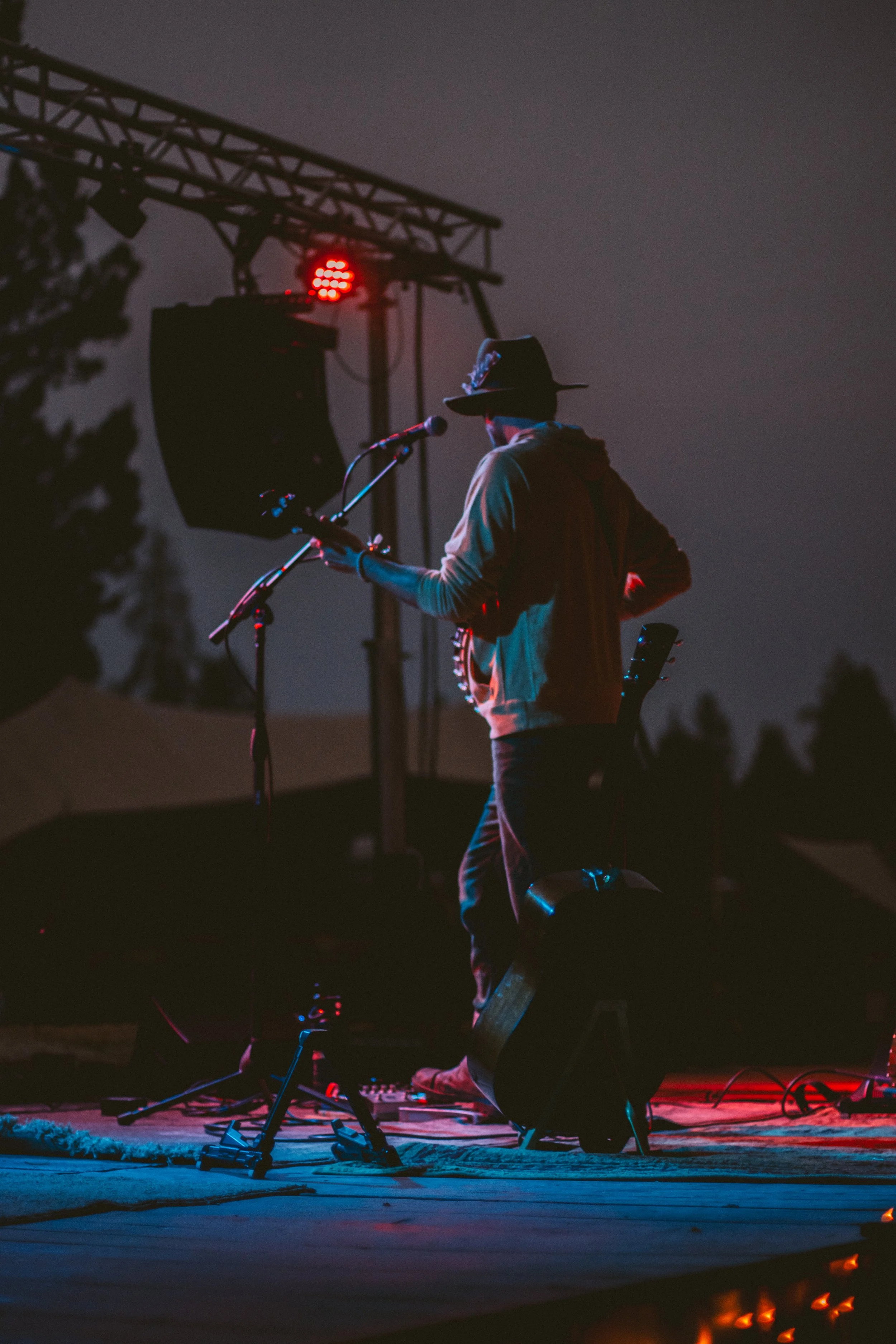 A person wearing a hat and hoodie, playing a guitar on an outdoor stage at night, with stage lights and equipment around.