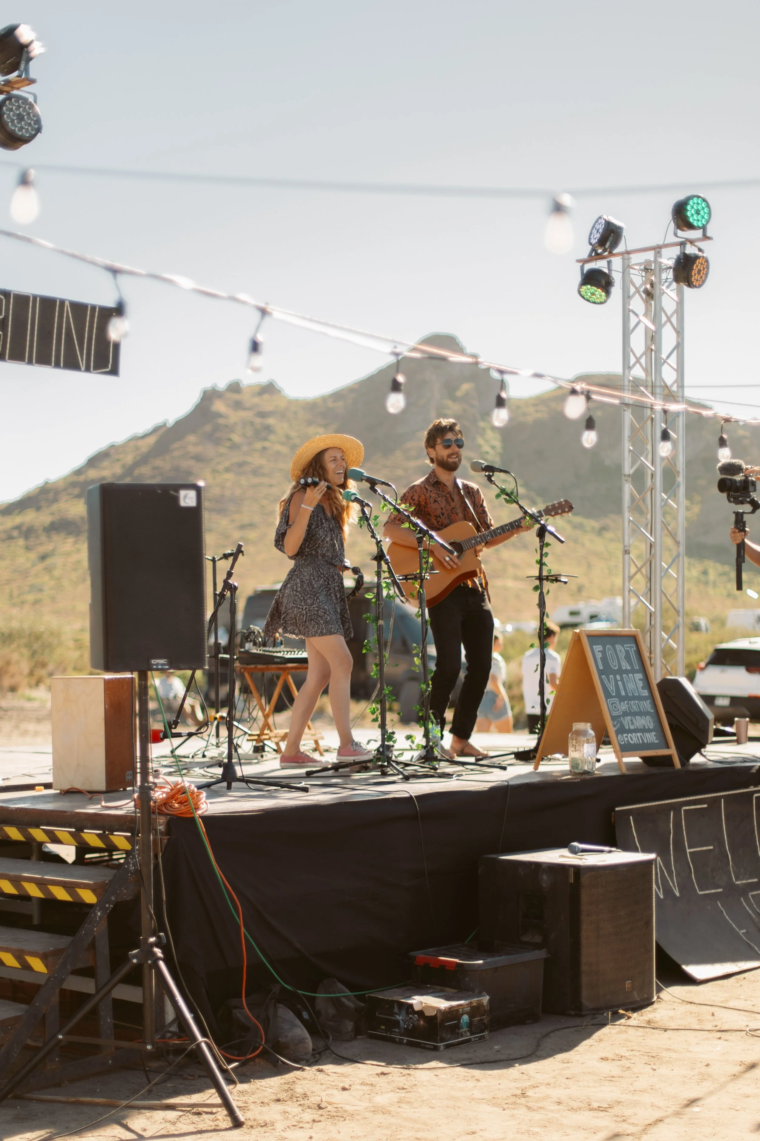Two musicians performing on an outdoor stage in a desert setting with mountains in the background. One is singing into a microphone while the other plays an acoustic guitar. Stage equipment, lighting, and a sign that reads "Fort Vine" are visible.