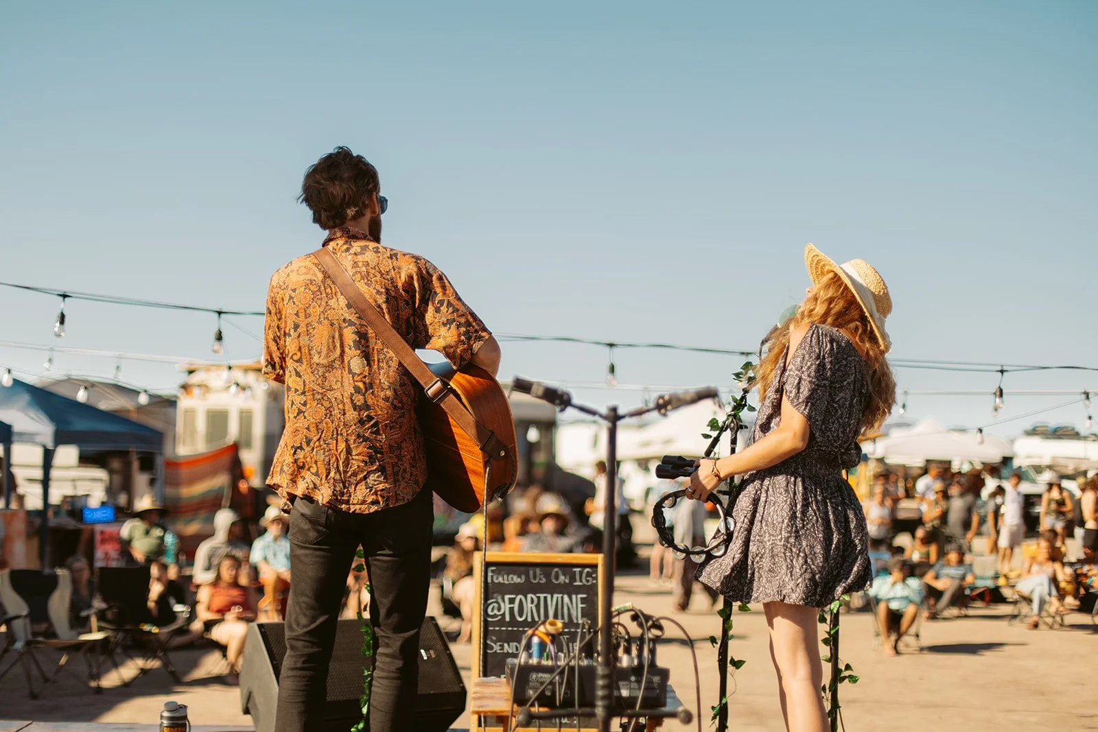 Two street musicians, a man with a guitar and a woman with long hair and a wide-brimmed hat, perform at an outdoor event on a sunny day, with a crowd and market stalls in the background.