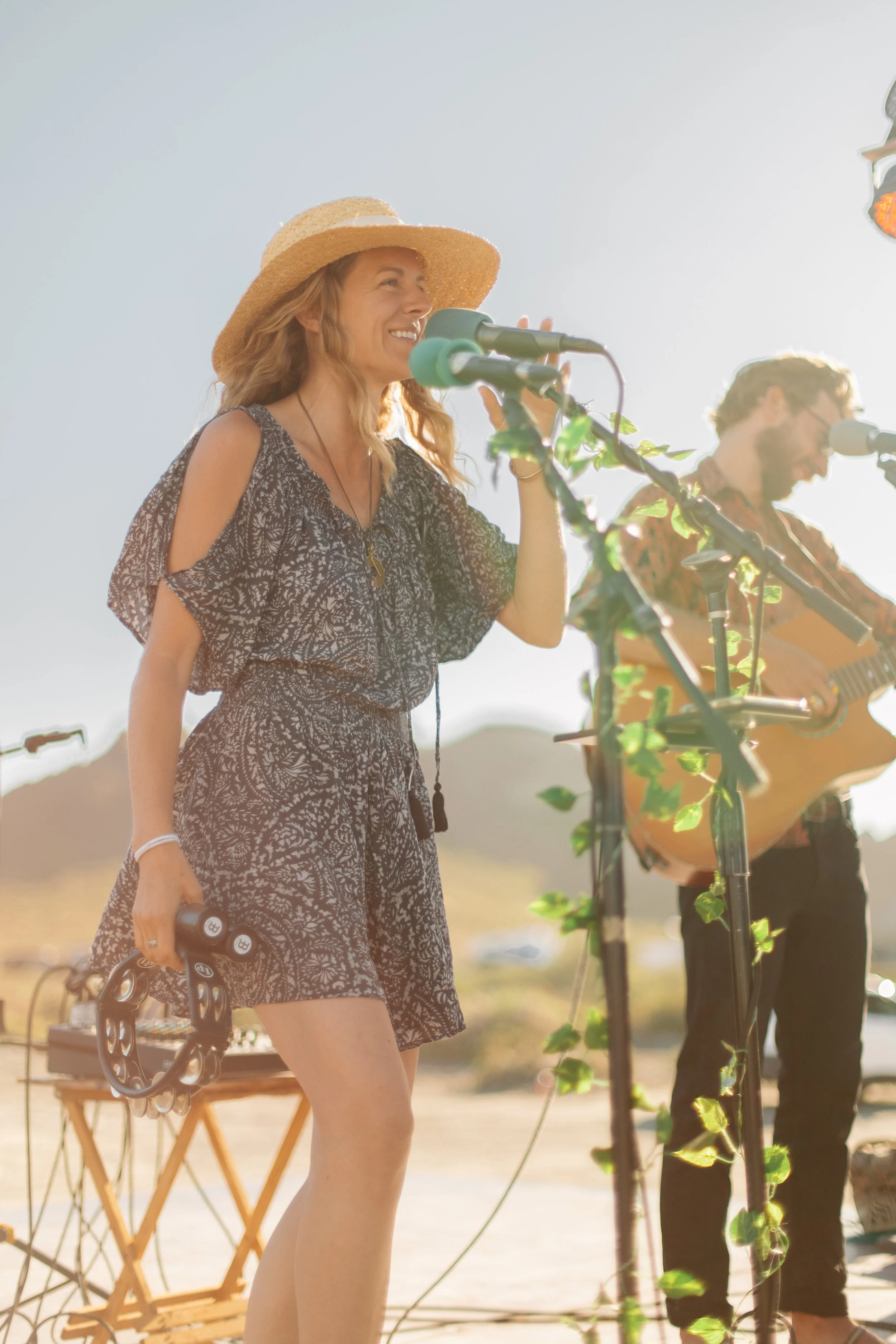 A woman in a black patterned dress, wearing a straw hat, holding a tambourine in one hand and standing in front of a microphone at an outdoor music event, with a man playing guitar in the background.