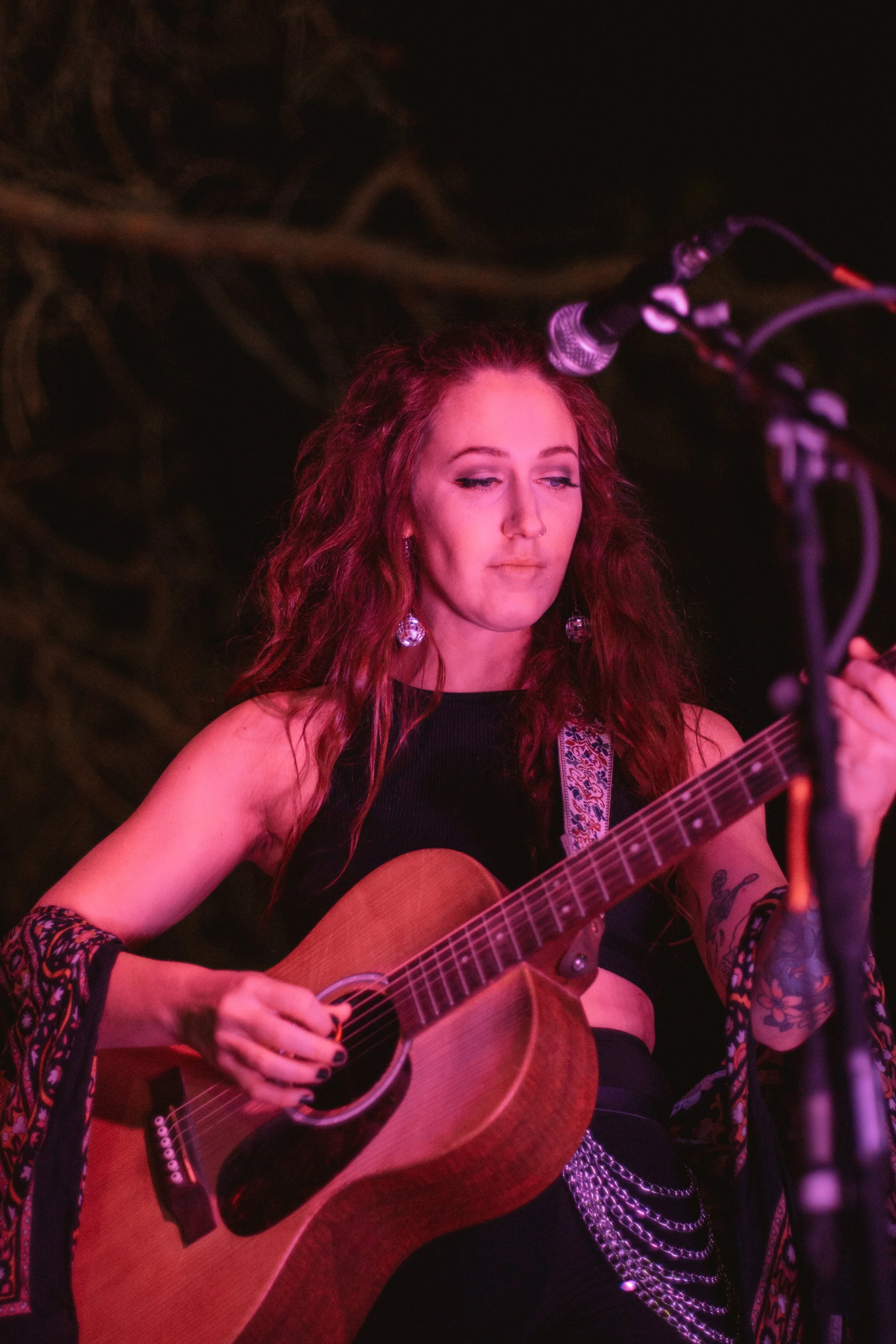 A woman with long, curly red hair and tattoos playing an acoustic guitar on stage under pink lighting, with a microphone in front of her.