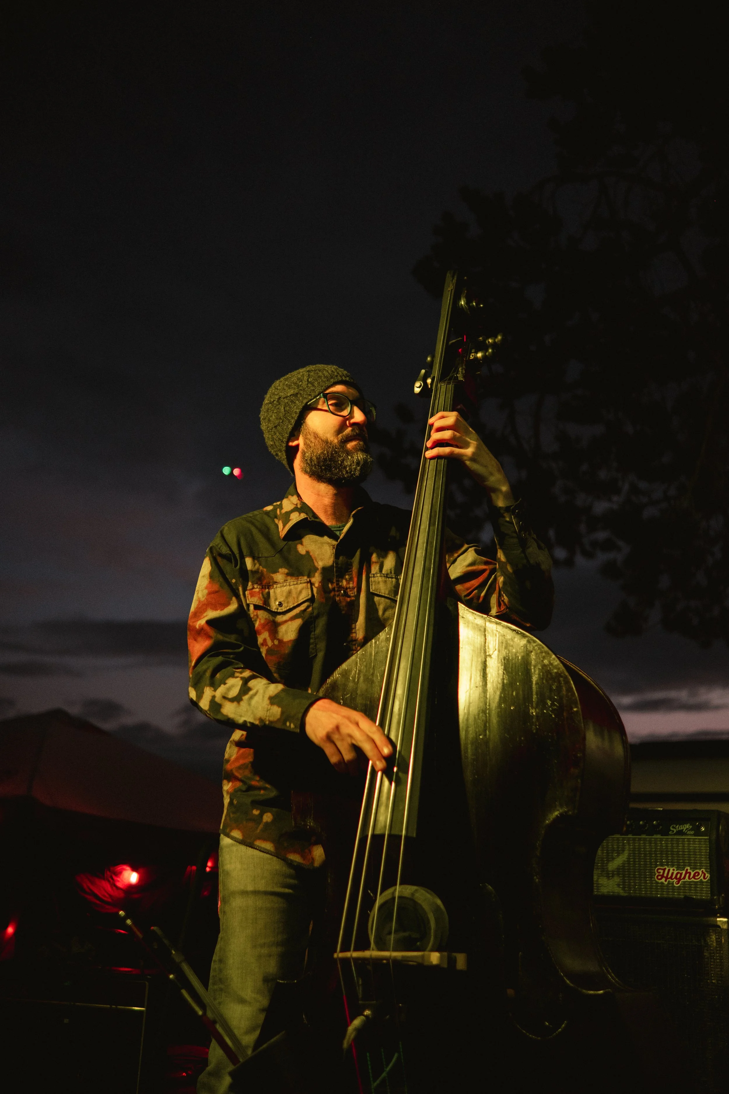 A man with glasses and a beard wearing a hat and camouflage shirt playing a double bass outdoors at night.