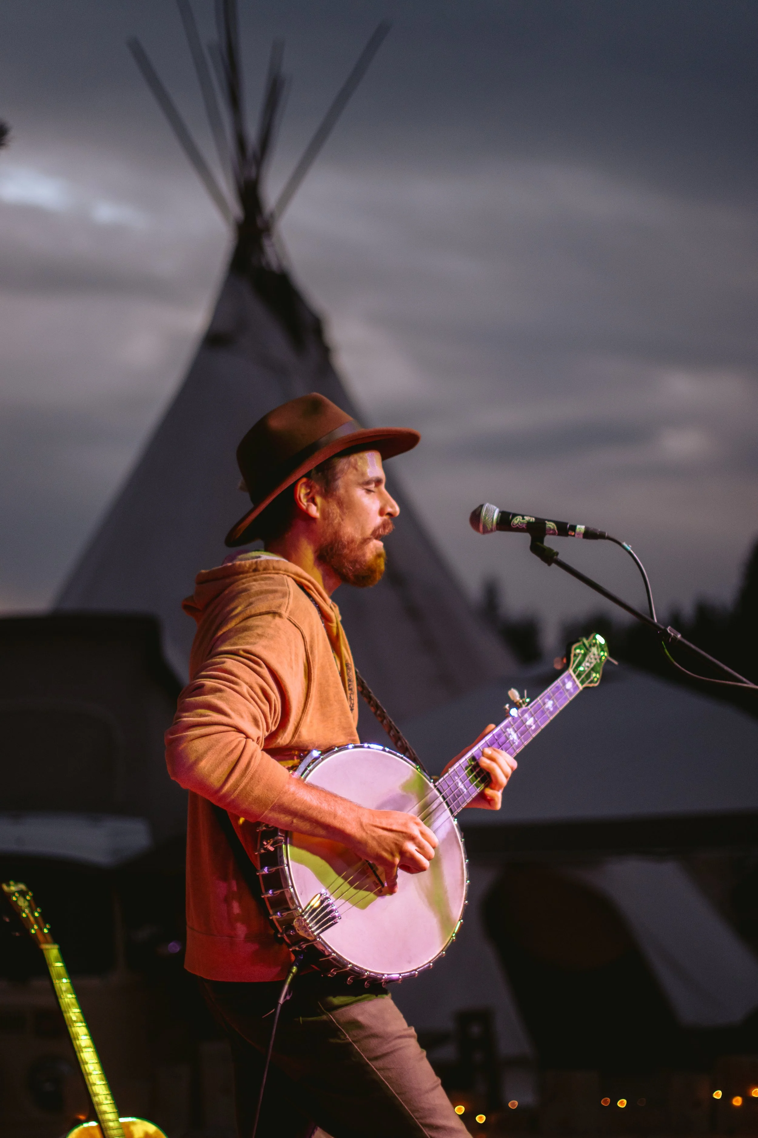 Man playing banjo and singing into microphone at an outdoor event, with large teepee in the background.