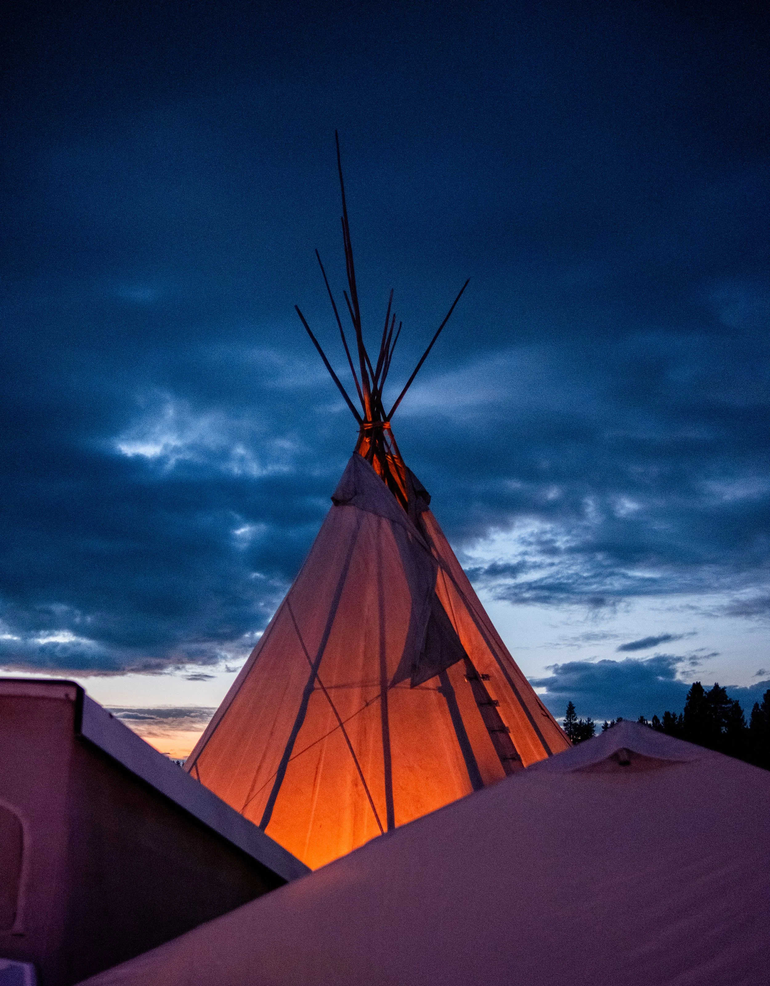 A traditional teepee tent illuminated from within against a darkening sky with clouds.