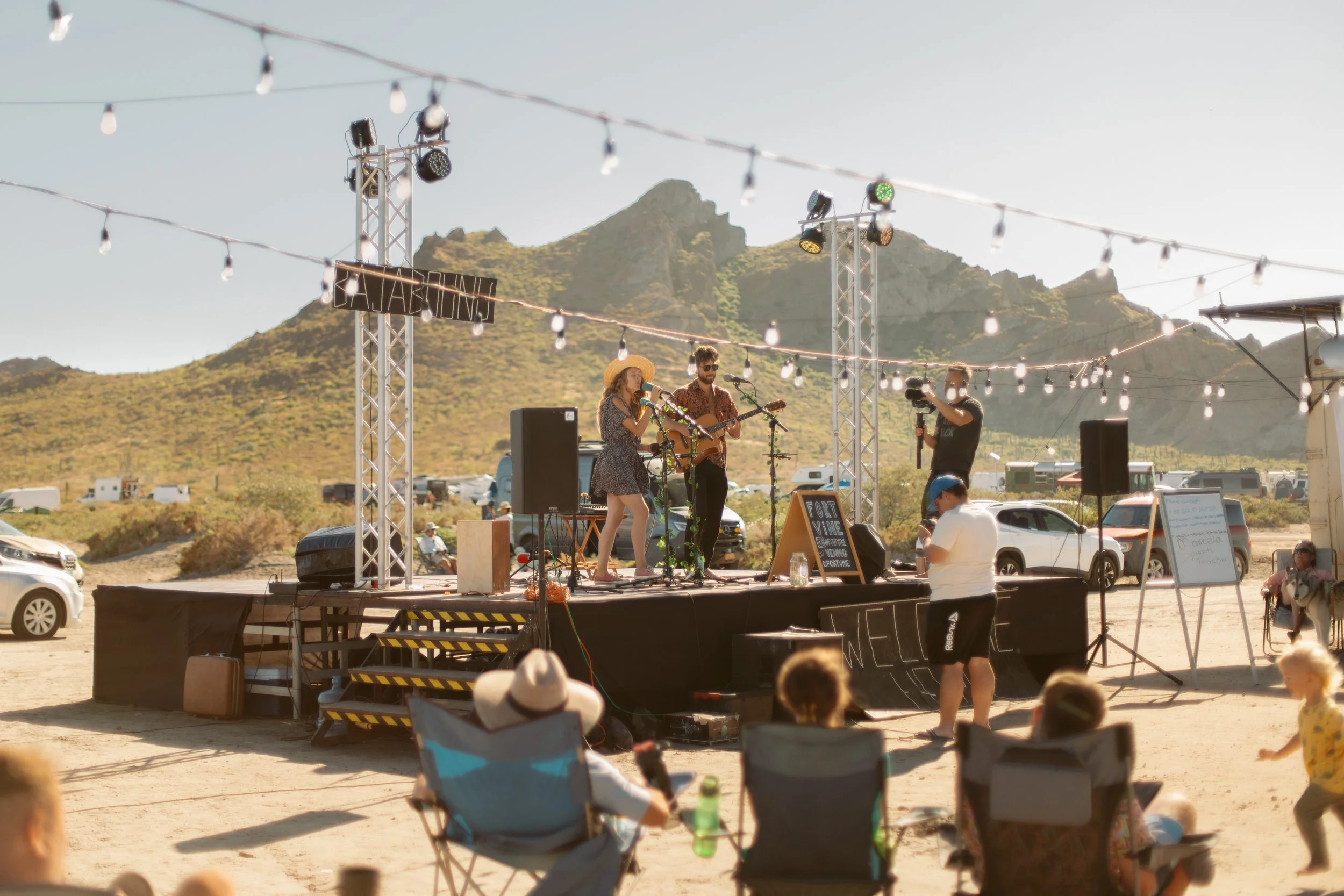 Live outdoor performance on a stage set up in a desert with mountains in the background, string lights overhead, and seated audience.