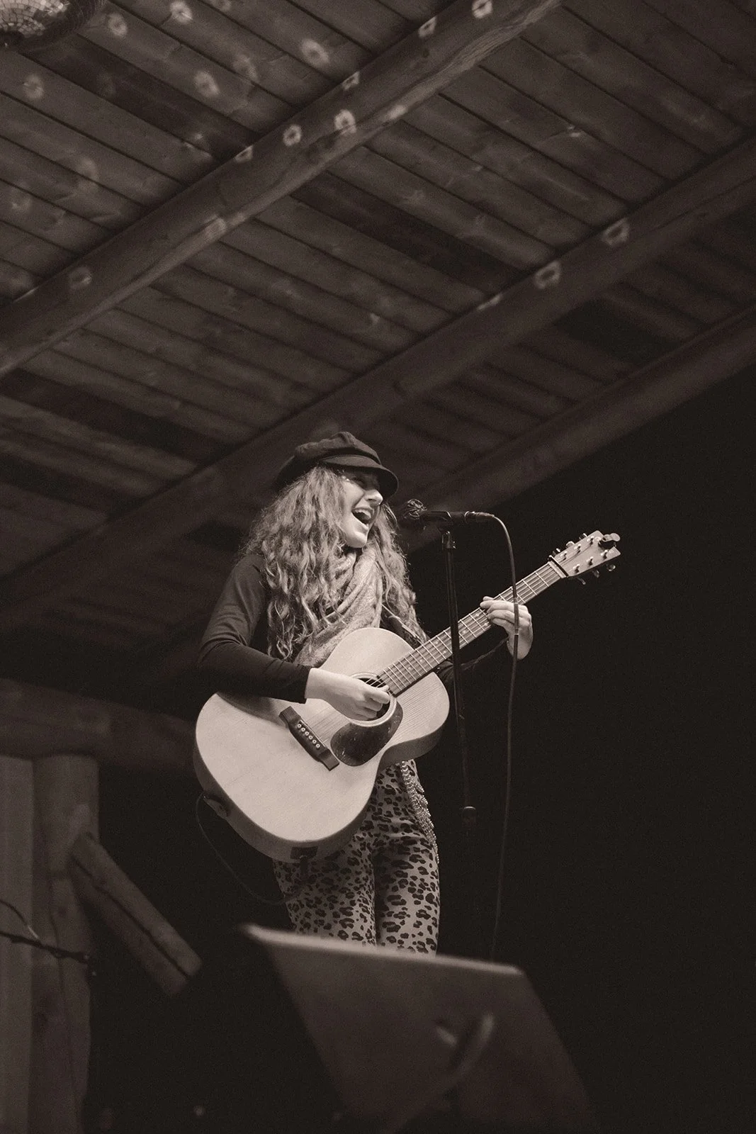 A woman with long curly hair, wearing a hat and leopard-print pants, performs on stage with an acoustic guitar, singing into a microphone.