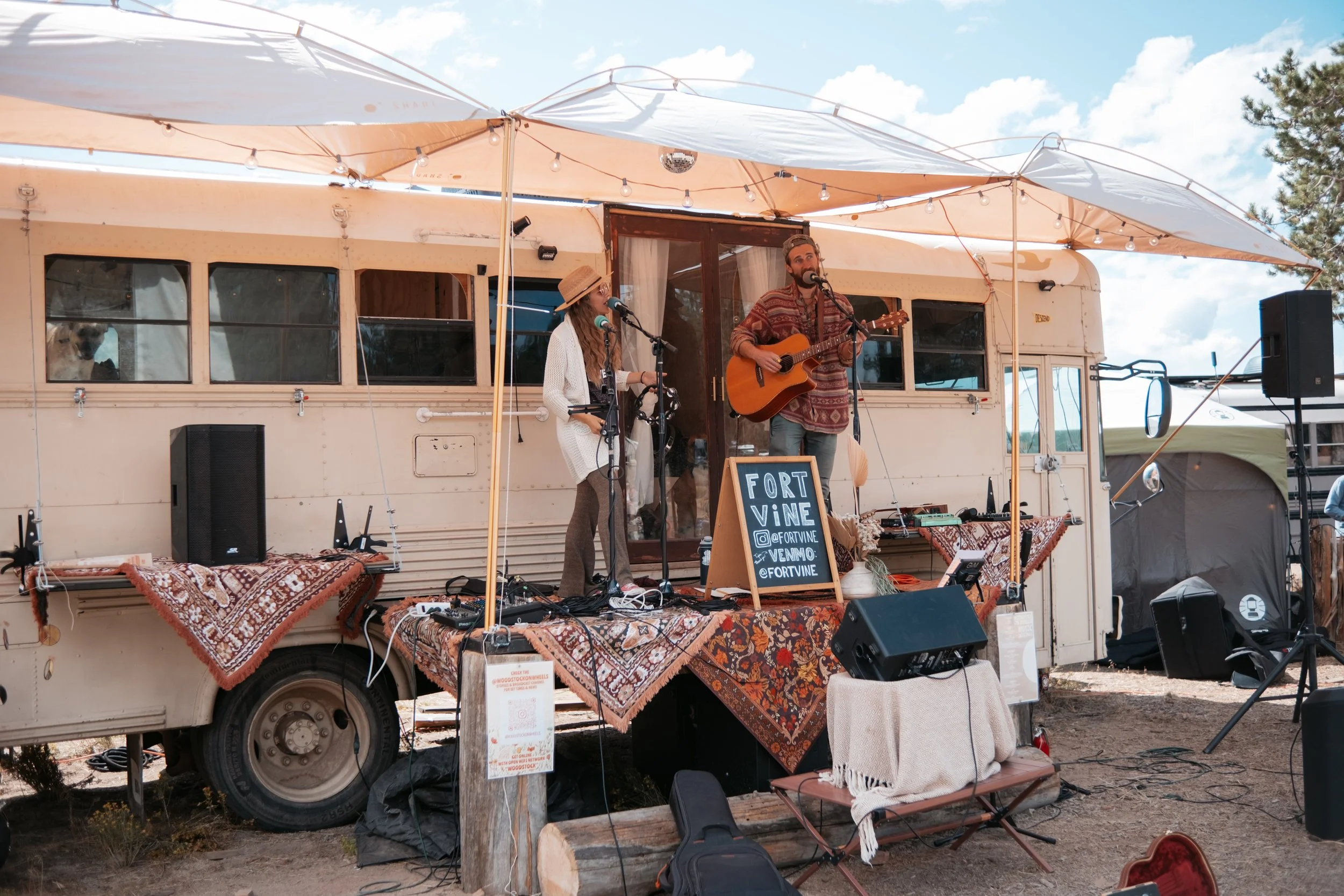 Musicians performing on a stage set up inside a vintage camper converted to a stage, outdoors under a partly cloudy sky. The man is playing an acoustic guitar and singing into a microphone; the woman is standing nearby with a microphone and a tambour