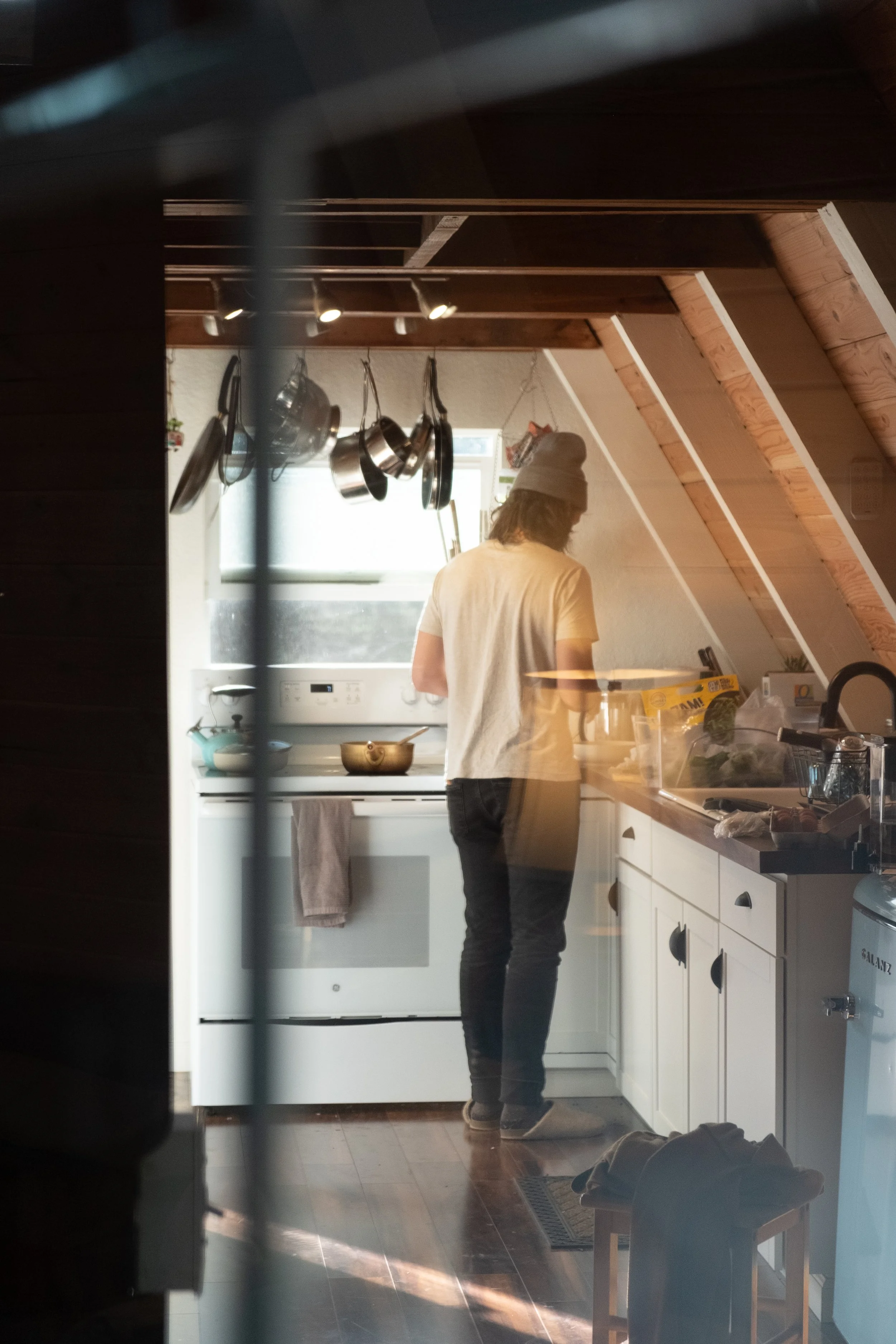 A person standing in a cozy kitchen with slanted wooden ceiling beams, looking away from the camera, surrounded by hanging pots and pans, kitchen appliances, and food items.