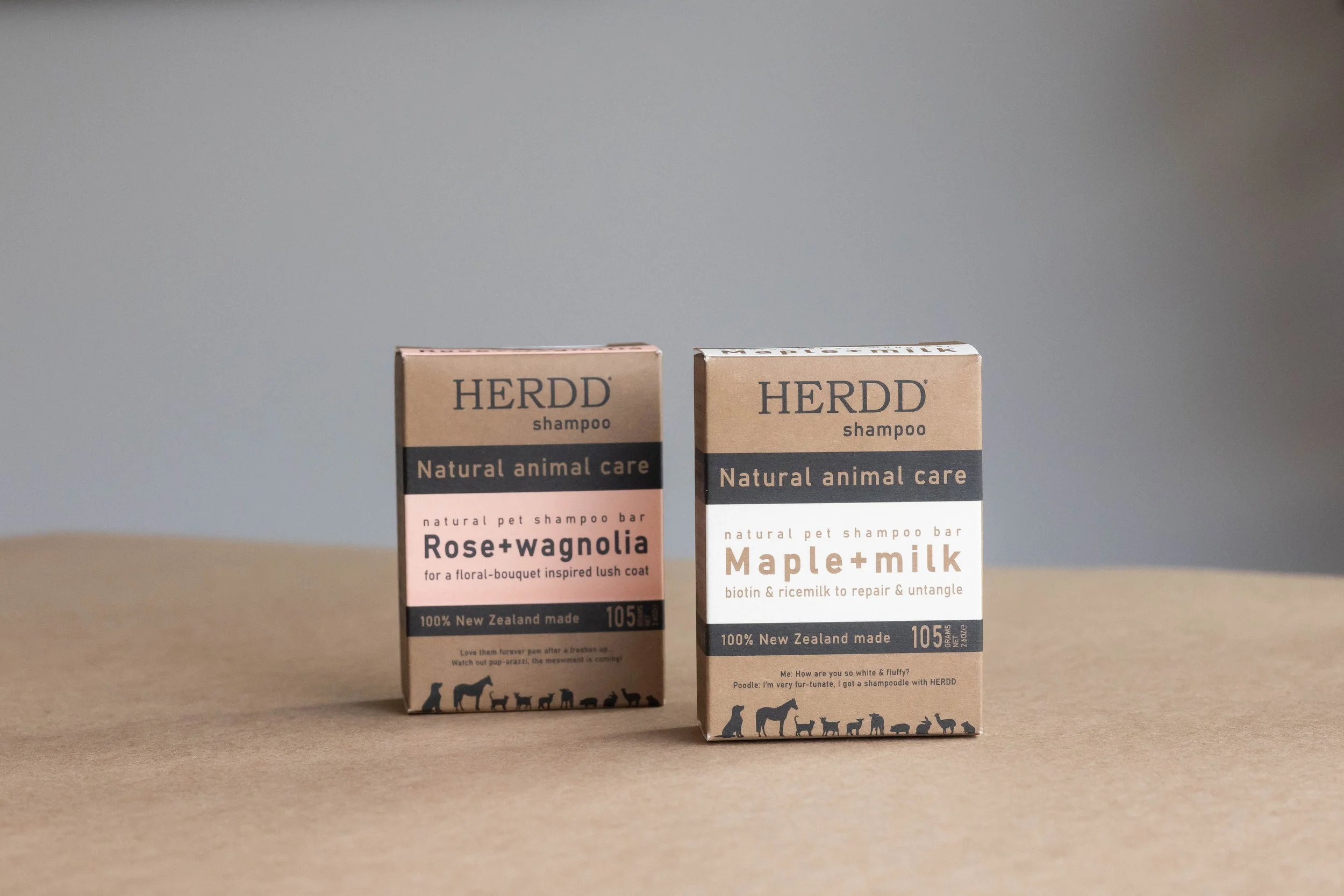 Two packages of HERDD shampoo bars on a beige surface with a gray background. One labeled 'Rose+wagnolia' and the other 'Maple+milk', both featuring black silhouettes of various animals at the bottom.