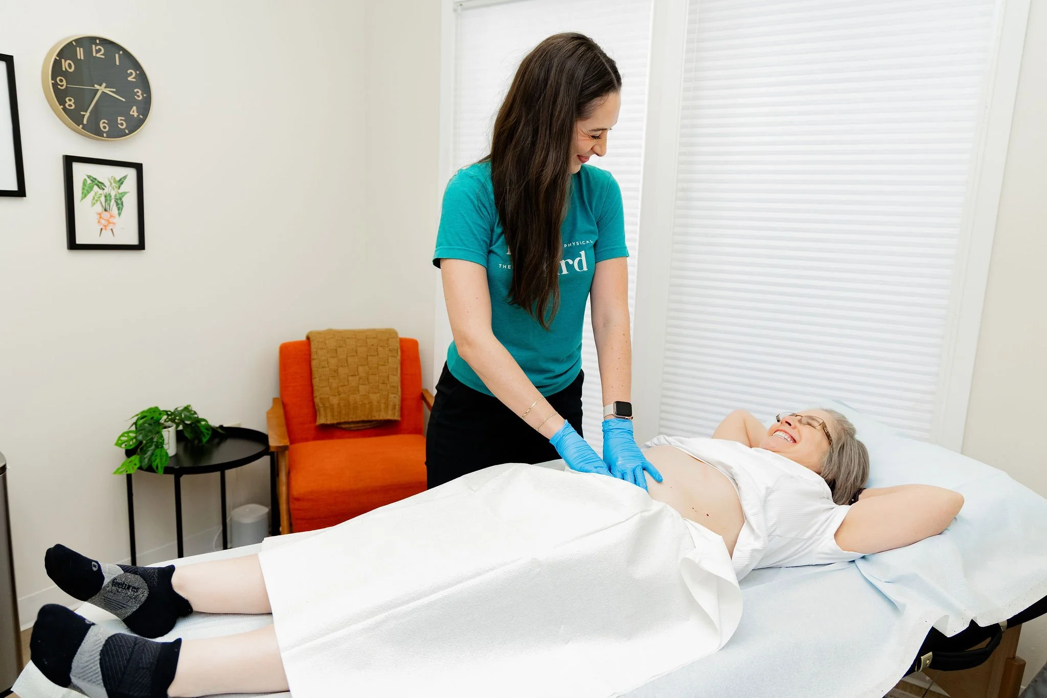 A healthcare professional, wearing blue gloves, is tending to an elderly woman lying on a hospital bed. The woman is smiling and wearing glasses, with her arms behind her head. The room has white walls, a round wall clock, framed pictures, a small black table with a potted plant, and an orange chair.