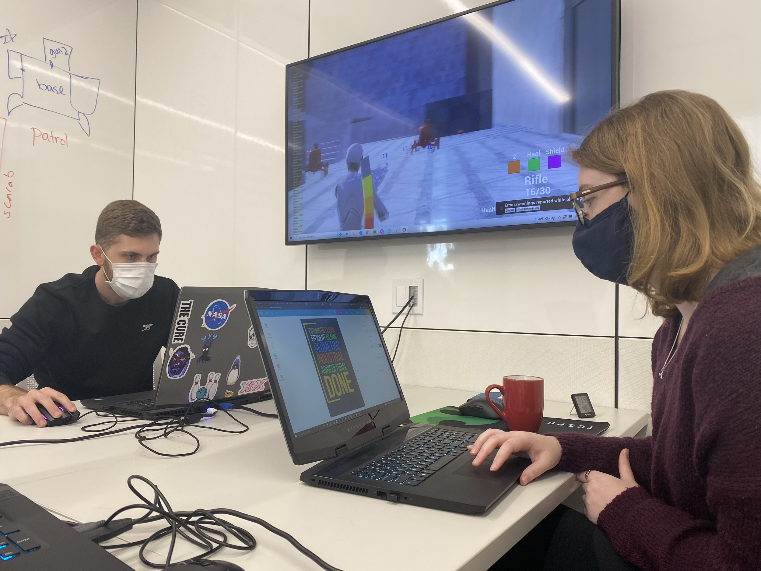 Photo of Dakota and Dube on their laptops. On a wall-mounted TV is AGRYOS: Recovering Eden.
