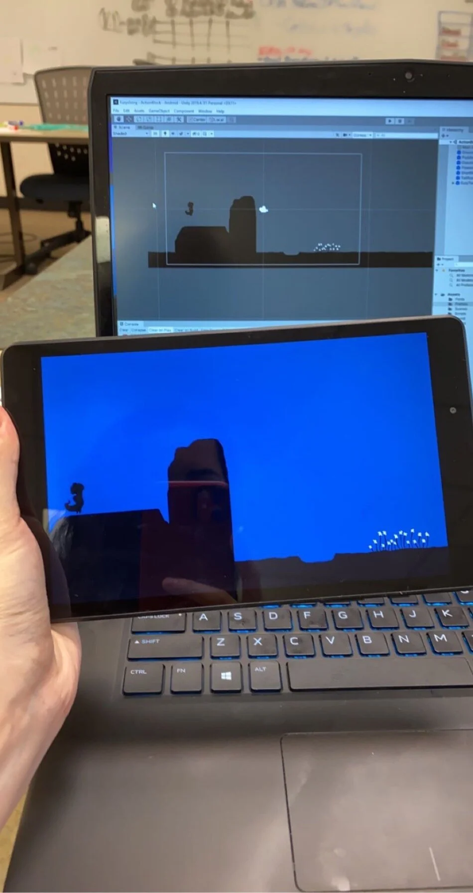 A photograph of someone holding a tablet in front of a laptop. The tablet and laptop both show the same thing: a small woman standing on the edge of a cliff, with flowers on the other side.