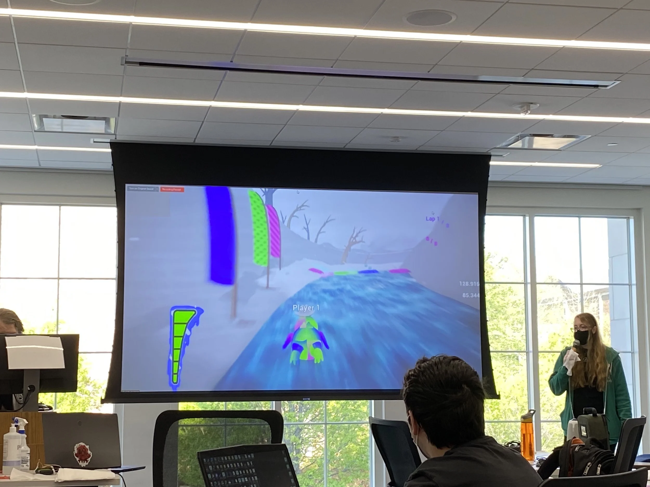 Photograph of Dube holding a microphone, giving a presentation. On screen beside her, a multi-colored penguin races down an icy track.