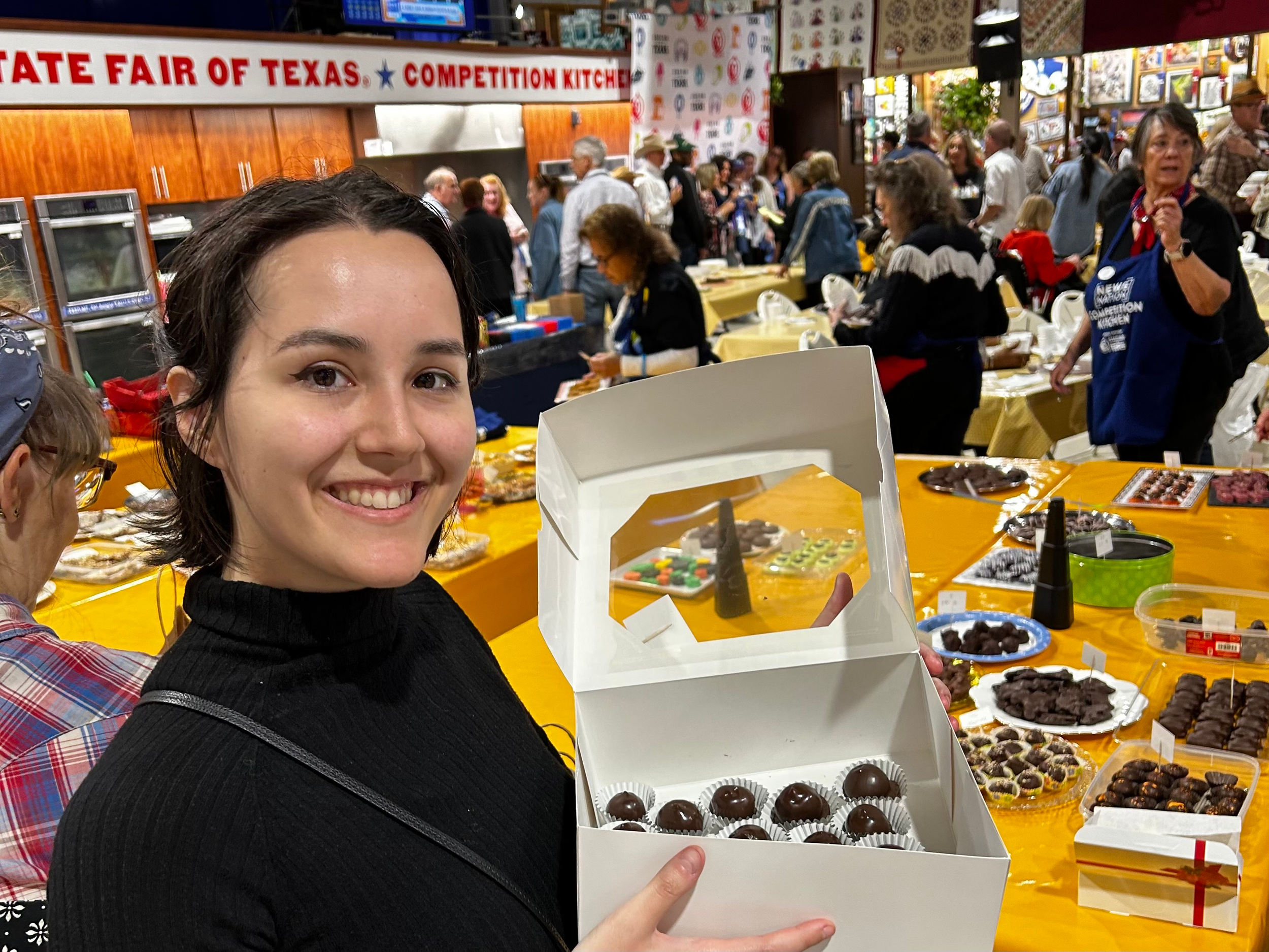 Nooka holds up a box of home-made chocolates at the State Fair of Texas Competition Kitchen. Behind her are tables filled witih other chocolate entries.