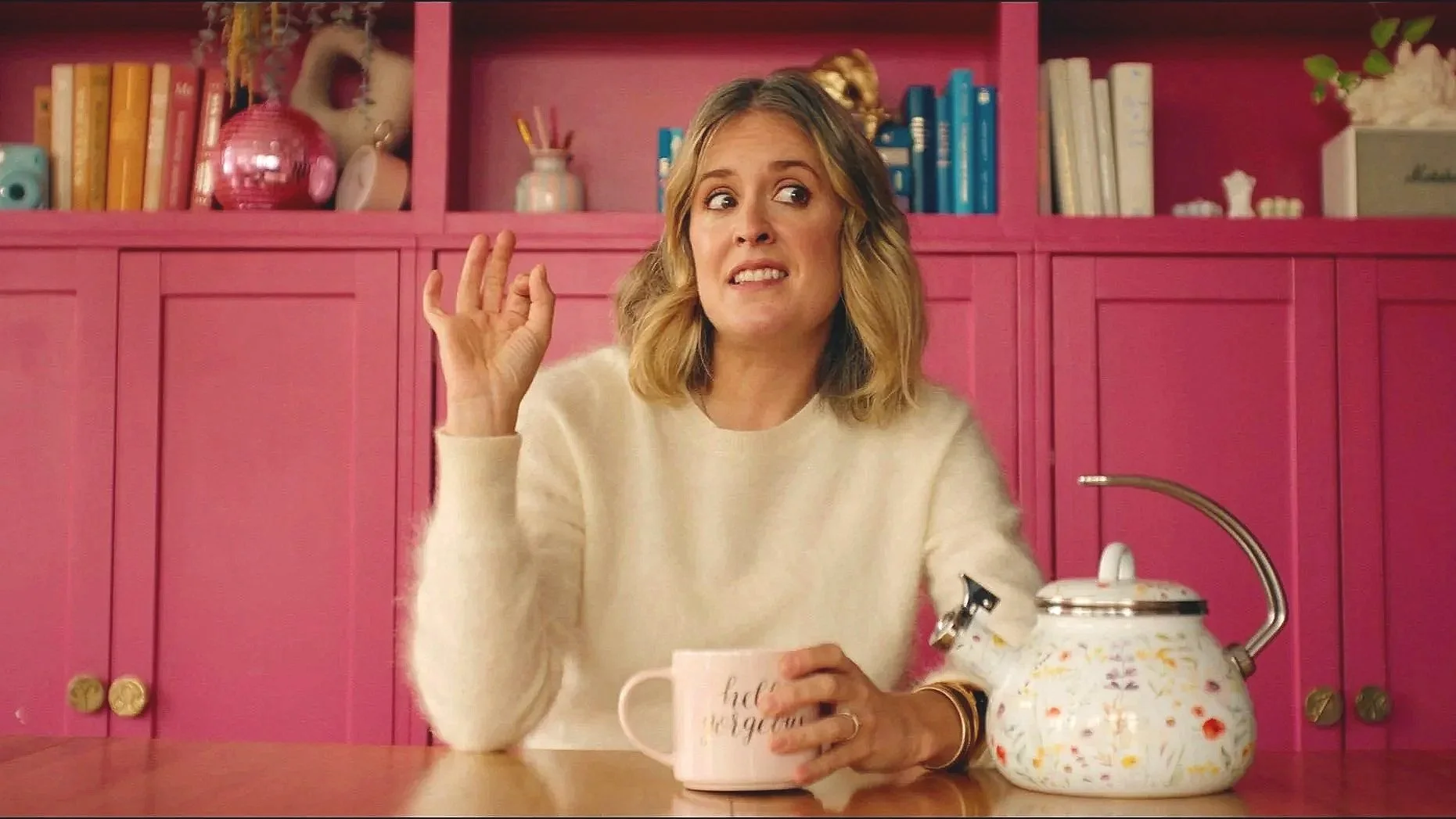 A social media influencer woman with shoulder-length blonde hair, wearing a white sweater, sitting at a kitchen table with a floral teapot, a pink mug, and a pink cabinet behind her. She appears to be surprised or confused.