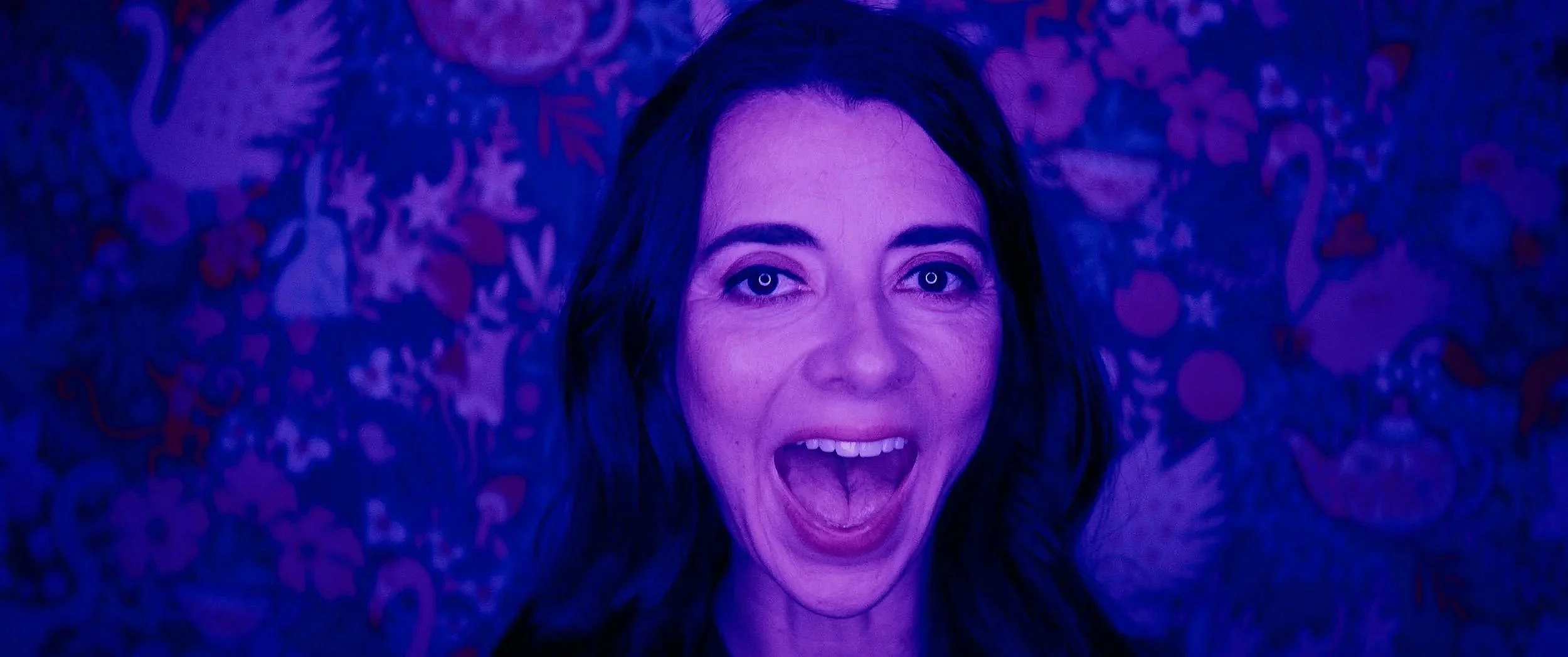 A still frame of Carmen Lynch, a woman with long dark hair smiling with her mouth open, in front of a colorful patterned background, illuminated with purple lighting.