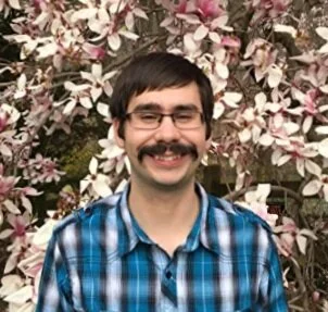 A man with glasses and a mustache smiling in front of blooming pink and white flowers, wearing a blue plaid shirt.