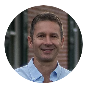 Portrait of a man with short brown hair wearing a light blue collared shirt, smiling outdoors with a blurred background.